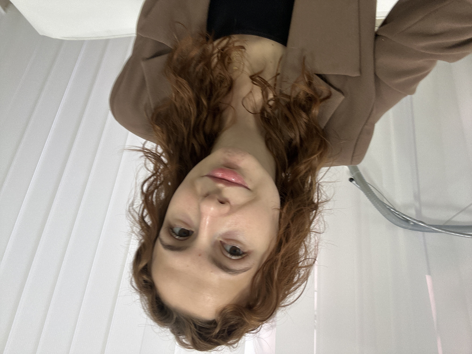 A medium close-up shot captures a young woman indoors in Novi Sad, Serbia, with the image rotated clockwise by 90 degrees. She has long, wavy reddish-brown hair that frames her face. She is looking directly at the camera with a calm, neutral expression and slightly glossy, pink lips. She wears a dark top, possibly black, underneath a light brown or beige blazer or jacket.
The background consists primarily of vertical white or off-white sheer curtains or blinds, suggesting a window or a softly lit indoor environment. To the upper right (when oriented correctly), a curved metallic object, possibly part of a chair or room fixture, is partially visible. The lighting is bright and even, highlighting her features. No visible text is present in the image.