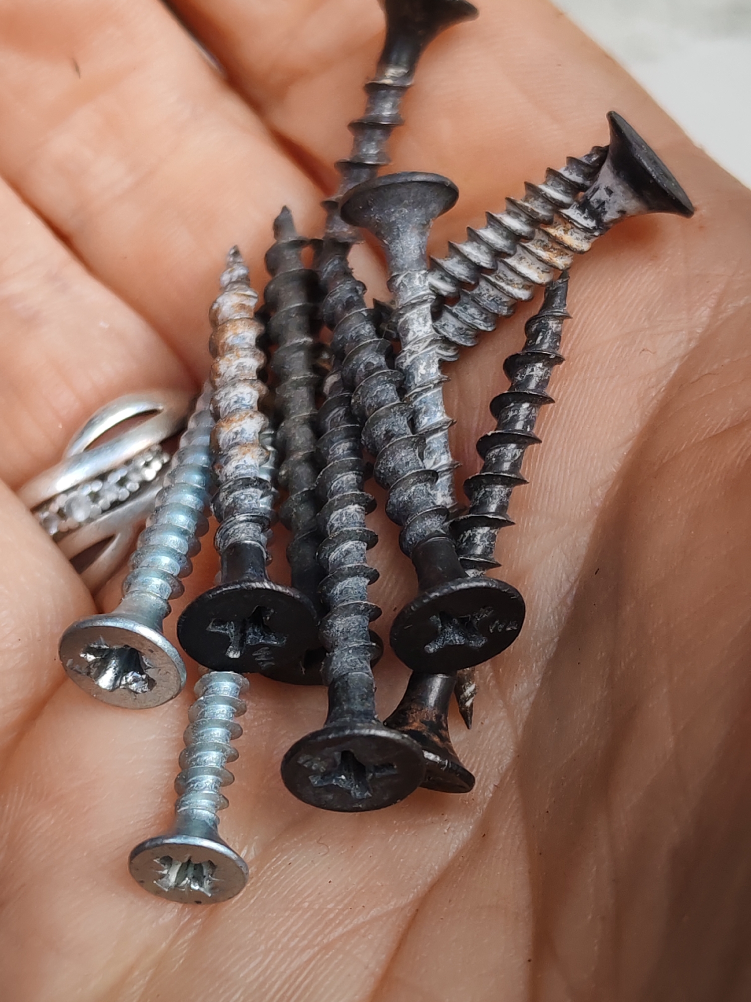 A close-up image shows a hand holding several screws. The screws are a mix of colors, some are silver and others are black. The silver screws appear to be newer than the black screws, which show signs of wear and oxidation. A silver ring is on the finger. The setting appears to be indoors. The location is Unknown, Unknown.