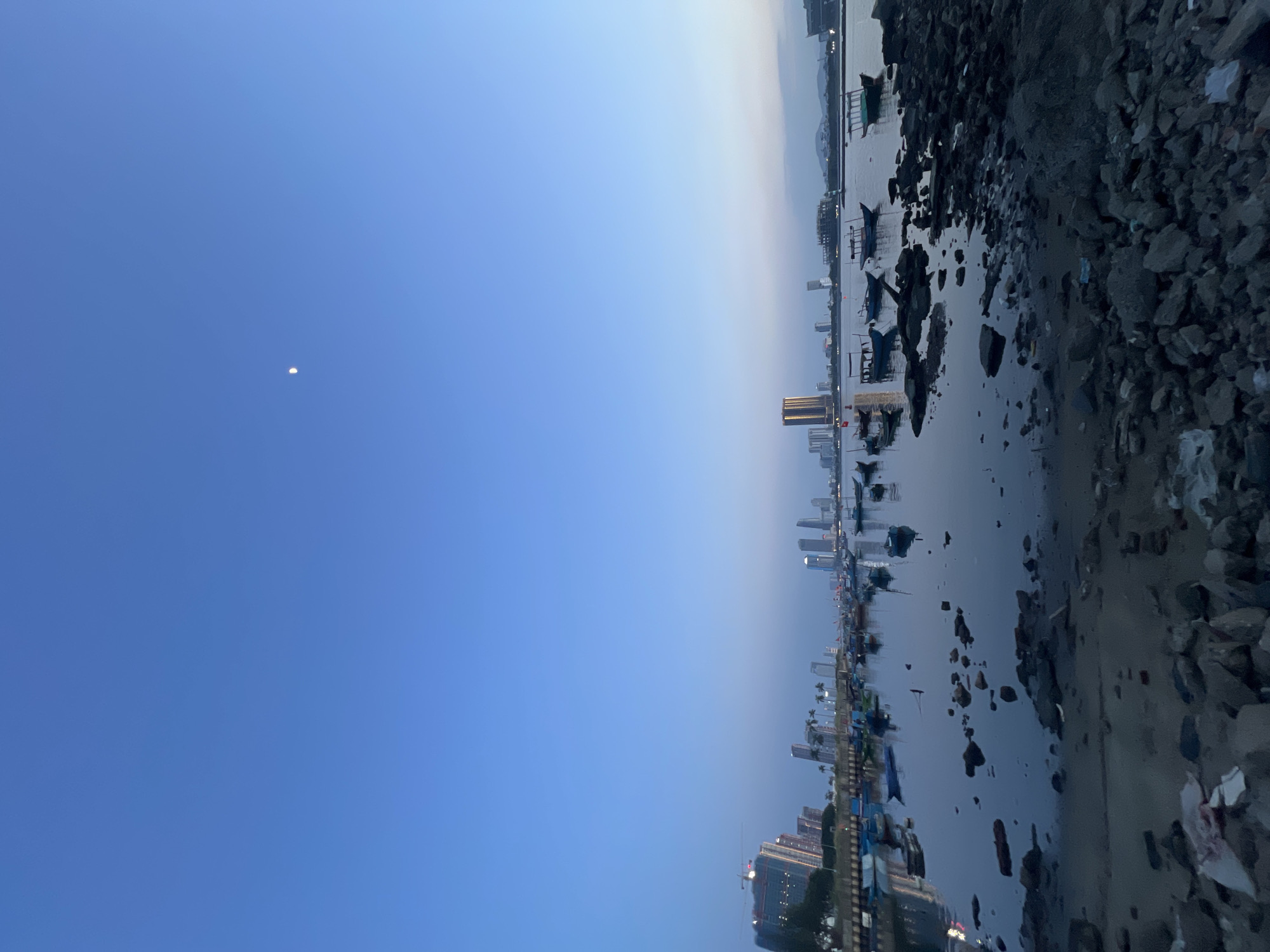 The image depicts a tranquil harbor scene at dusk in Da Nang, Vietnam. The sky is a clear, deep blue, transitioning to a lighter hue near the horizon, with a sliver of the moon visible. In the distance, the skyline of Da Nang is illuminated by city lights, featuring several modern skyscrapers and buildings. The foreground is dominated by a rocky shoreline and a calm body of water reflecting the sky and the city lights. Numerous small fishing boats are moored in the water, some with canopies. The overall atmosphere is serene and peaceful, capturing the quiet beauty of the city as day turns into night.