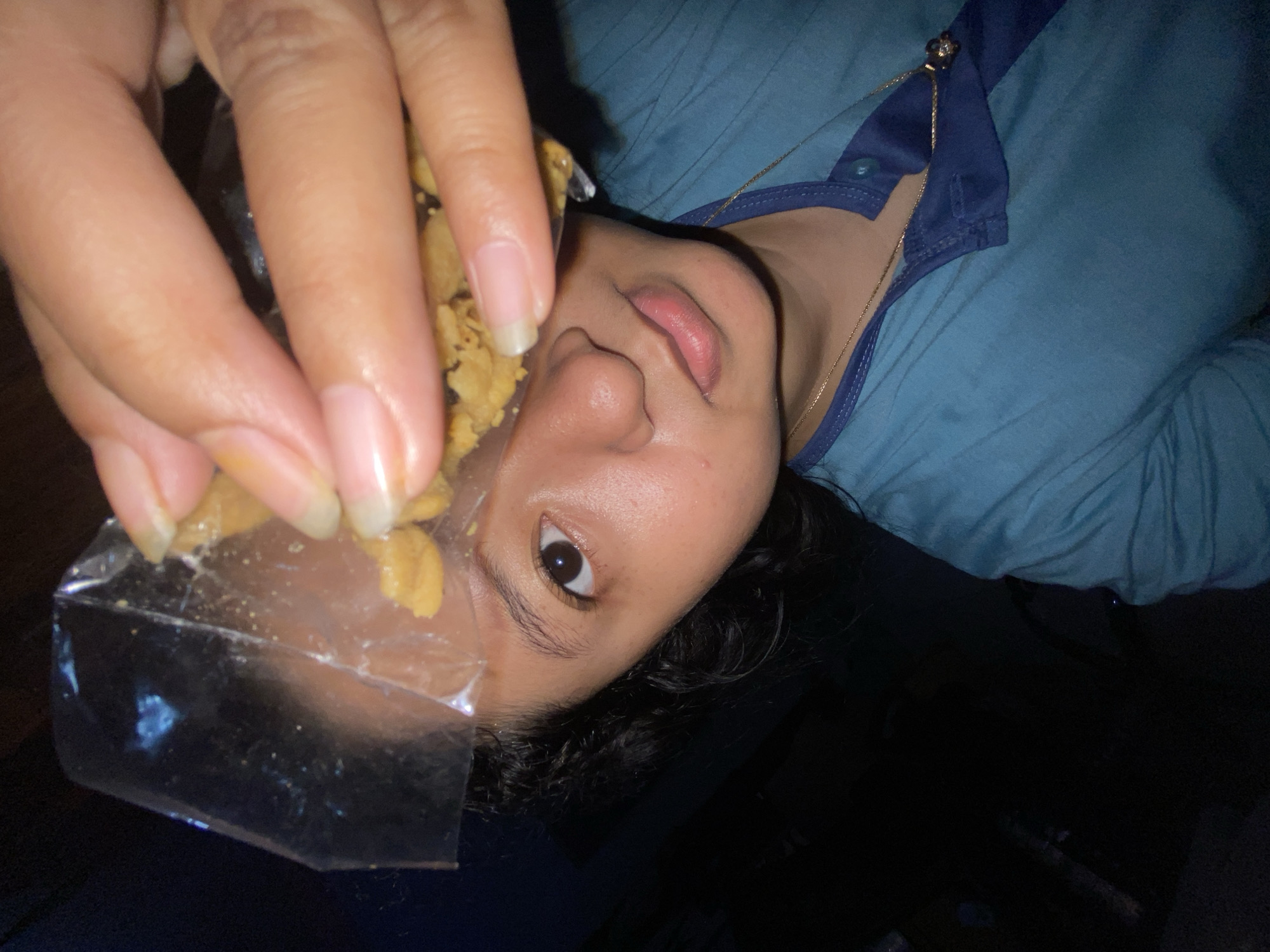 The media file shows a close-up, rotated view of a person's face, partially obscured by their hand holding a small clear bag of food. The person has dark hair, dark eyes, and is wearing a blue shirt with a darker blue collar. A thin gold chain necklace is visible on their neck. The food in the bag appears to be small, light-brown pieces, possibly nuts or snacks. The lighting suggests it could be indoors at night. The provided location context is Gelam, Indonesia, though no specific visual cues in the image confirm this. The activity depicted is the person holding food, possibly about to eat or share it. The image is focused on the person's face and hand, with the background being dark and out of focus. There is no visible text in the image.