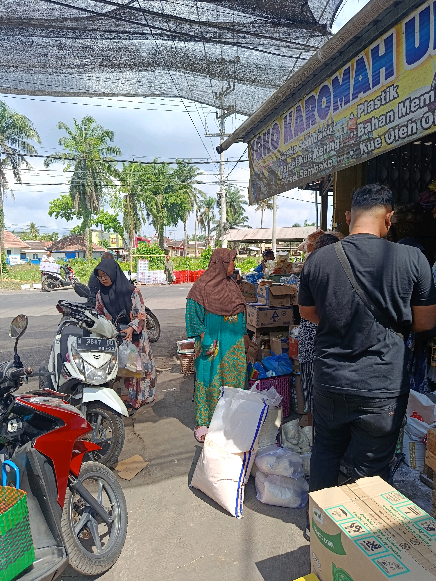 A street-level, outdoor scene in Jatiroto, Indonesia, captures a bustling small shop front under a woven sunshade. Palm trees and a cloudy sky are visible in the background. In the foreground, several motorcycles are parked on a concrete path. A woman in a black hijab and patterned dress is attending to a white motorcycle, holding a plastic bag of purchases. Next to her, a woman in a teal and brown hijab wears a patterned dress and stands near a display of goods. Further back, a man in a black t-shirt and dark pants stands facing away from the camera, appearing to interact with the shop. The shop itself is laden with various products, including boxes labeled with text, sacks of rice, and baskets. A sign above the shop reads "TOKO KAROMAH" and lists items like "Aneka Sembako" (various groceries), "Plastik" (plastic), "Bahan Memasak" (cooking ingredients), "Pariel Kue Kering" (dried cake parcels), and "Kue Oleh Oleh" (souvenirs). A motorcycle with a license plate "N 3687 UD" is prominently displayed. The overall impression is one of a busy marketplace, likely a local grocery or convenience store catering to pedestrians and motorcyclists.