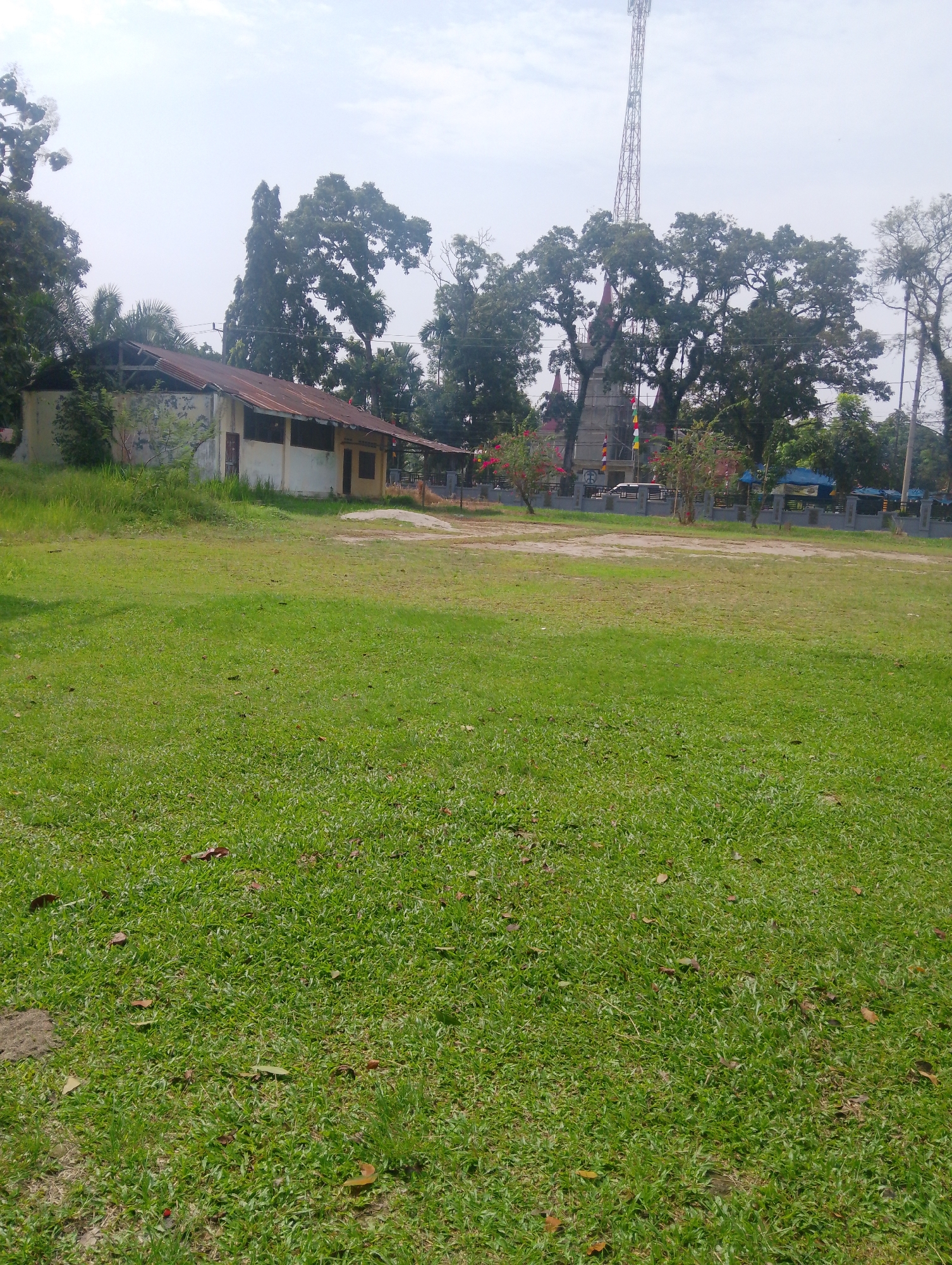 This image depicts an outdoor scene in Serbelawan, Indonesia. A wide expanse of green grass dominates the foreground, with scattered dry leaves on the ground. In the middle ground, a long, low building with a corrugated metal roof and light-colored walls stands to the left. The building appears to be partially dilapidated, with exposed roof beams. To the right of the building, a grey concrete fence encloses an area with some vegetation and structures, including what appears to be a tower in the background. Trees, some with dense foliage, are scattered throughout the scene, with a tall communication tower visible above the trees in the far background. The sky is overcast with a hazy, bright appearance. No people are visible in the image. The overall environment suggests a semi-rural or institutional setting.