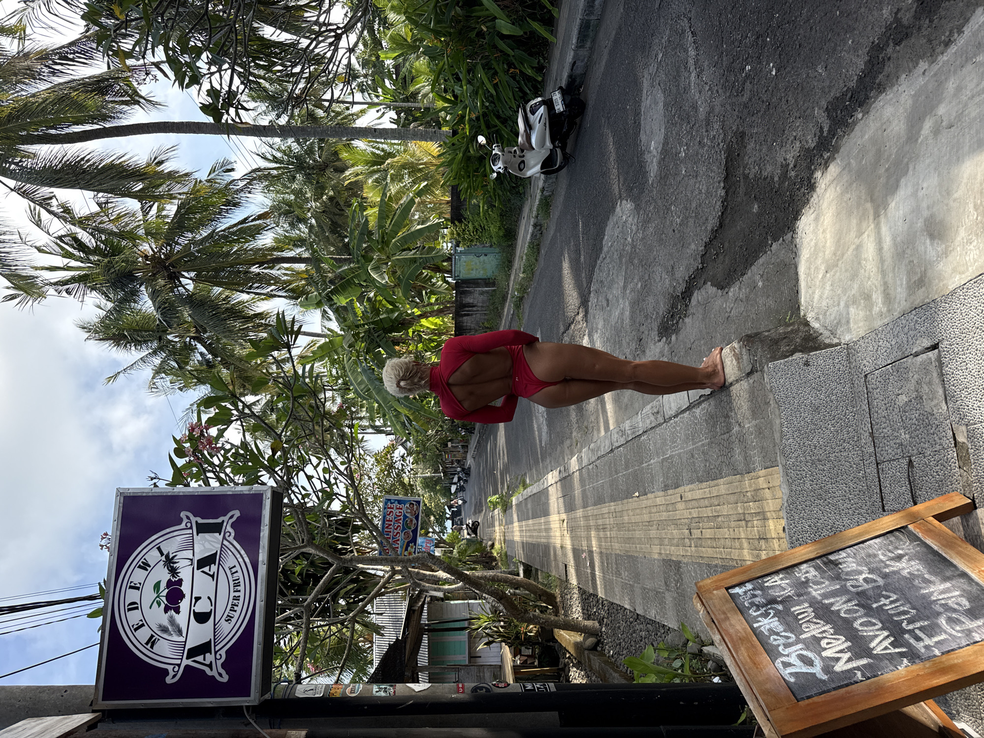 Here is a detailed description of the image:
**Scene:** This image shows a street scene in what appears to be a tropical or subtropical location, judging by the palm trees and lush vegetation.
**People:** A woman is standing on the sidewalk. She has blonde hair and is wearing a red one-piece swimsuit. Her back is to the camera.
**Objects:**
* A motorcycle is parked on the side of the road.
* Several signs are visible: one reads "MEDEW ACAI SUPER FRUTT", another reads "CHINESE MASSAGE", and a chalkboard advertises "Breakfast" with various food items listed (e.g., "Medewi Iced", "AVO ON TO", "Fruit Bo").
* Palm trees are growing along the road, adding to the tropical atmosphere.
**Actions/Interactions:** The woman is simply standing on the sidewalk, appearing to observe the scene. There are no obvious interactions between the woman and any other elements in the image.
**Environment:** The scene takes place on a paved road with a concrete sidewalk. The road is lined with lush greenery and businesses. The sky is partly cloudy.