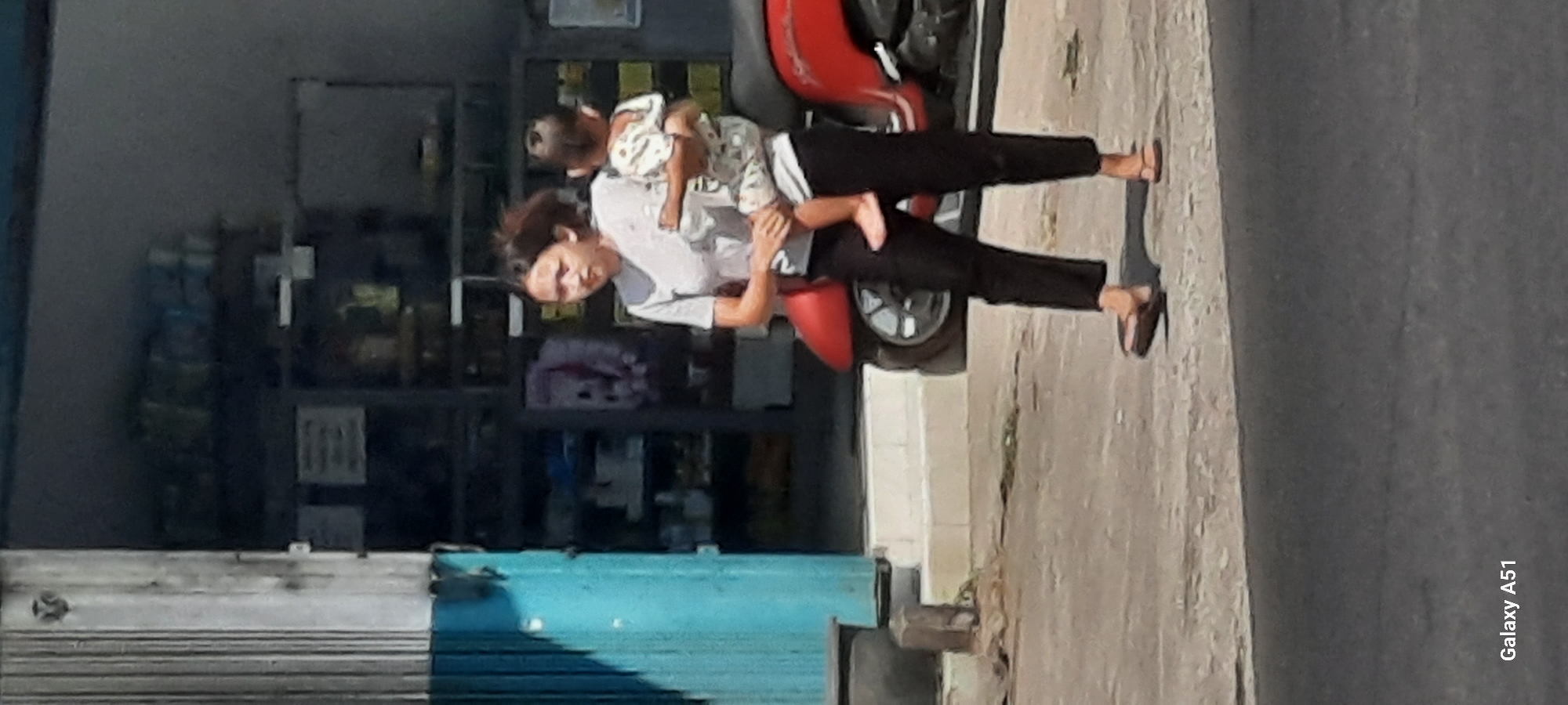 A young woman with dark hair is carrying a toddler. She is wearing a light-colored t-shirt and dark pants, and sandals. The toddler is wearing a patterned onesie. They are walking on a sidewalk in front of a store with a blue rolling door and shelves stocked with various items behind glass. A red scooter is parked to the right of the woman. The scene appears to be in Selopuro, Indonesia. The lighting suggests it is daytime.