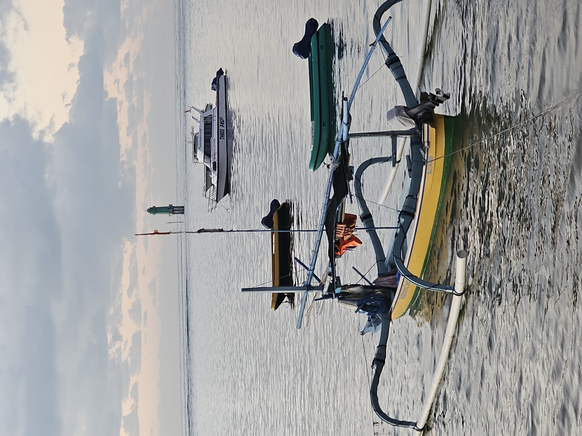 The image captures a serene coastal scene likely near Denpasar, Indonesia. In the foreground, a traditional outrigger boat with a yellow hull and green accent sits calmly in the water. Two green objects, possibly kayaks or buoys, are perched atop the boat. Behind it, further out in the water, a larger, white motorboat is visible. In the distance, a green structure, possibly a navigation marker or lighthouse, stands near the shore. The sky is overcast with a blend of gray and light peach tones, suggesting either dawn or dusk. The sea is relatively calm, with gentle ripples reflecting the ambient light.