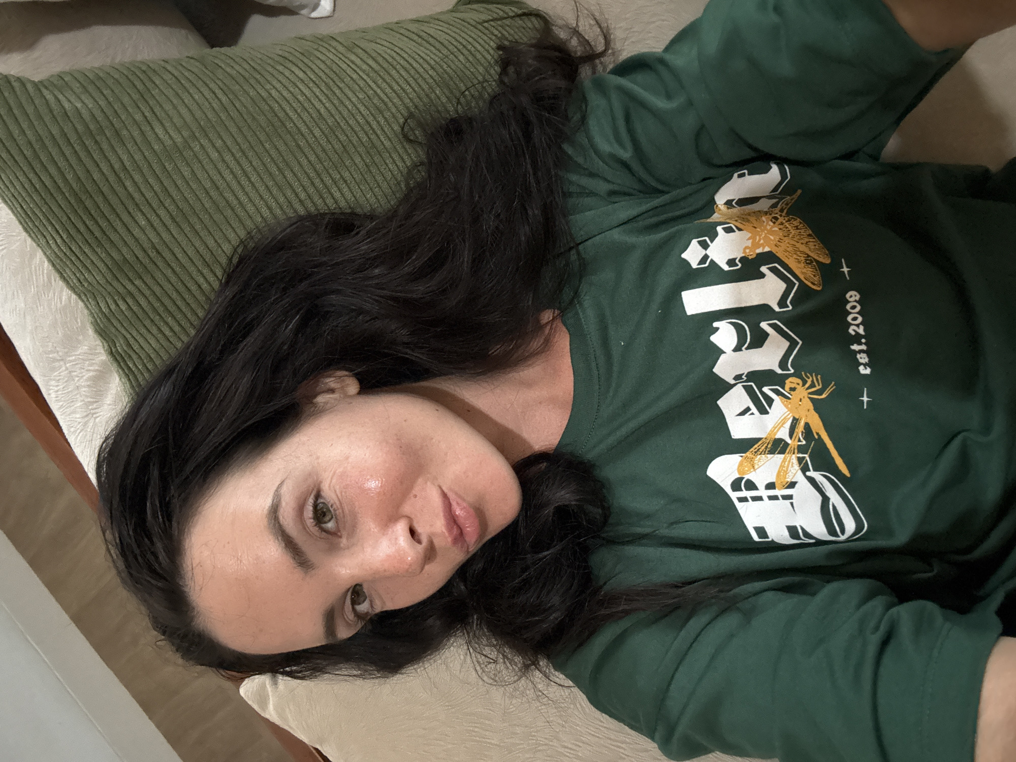 A woman with long, dark hair is lying down, looking at the camera. She is wearing a green long-sleeved shirt with a graphic design on it, which appears to say "Berlin" with two dragonfly graphics and "est. 2009". She is resting on a couch with a textured green pillow behind her. The background is blurred but appears to be an indoor setting.