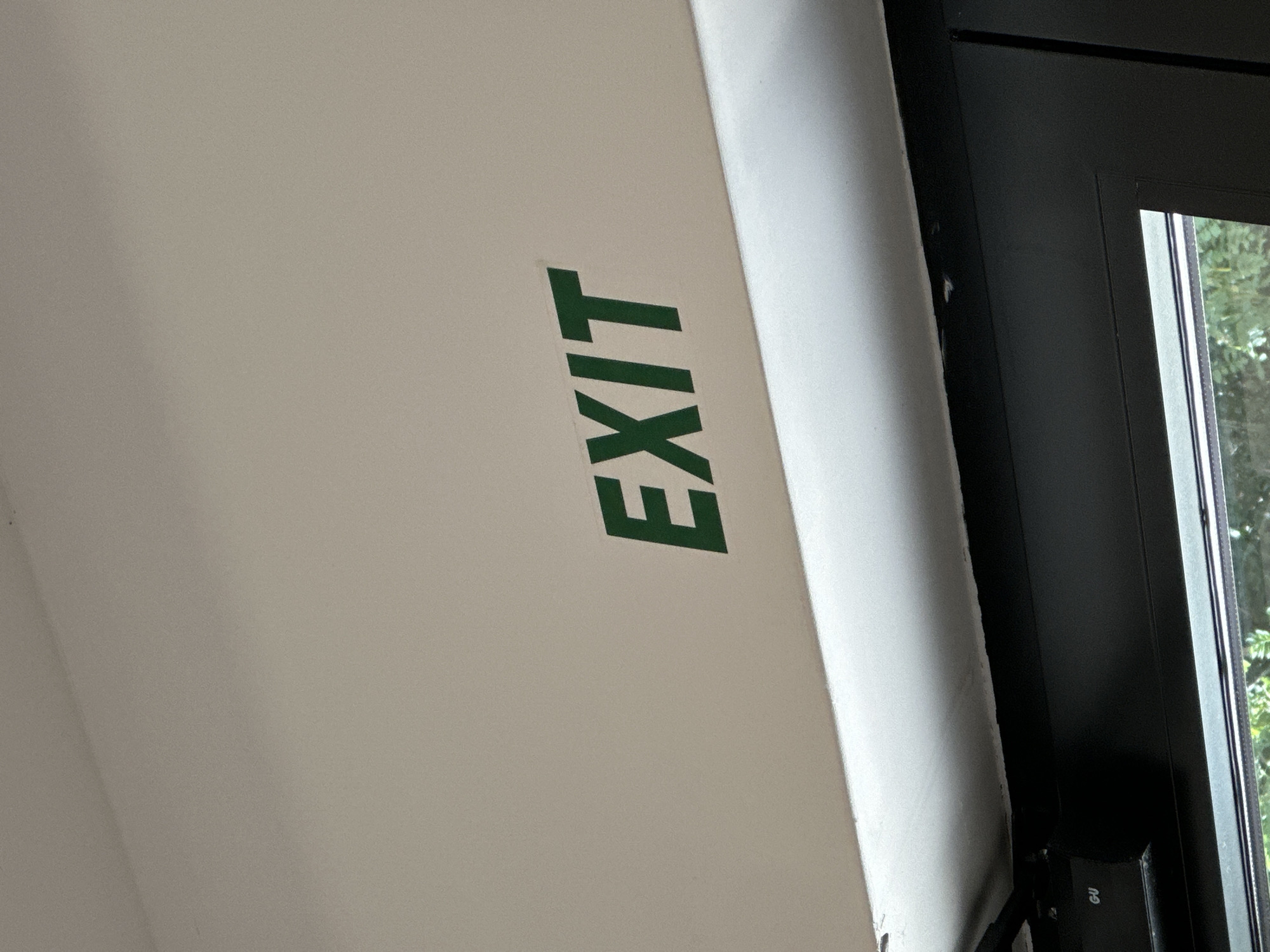 The image shows a close-up of a white ceiling with a green "EXIT" sign. Below the ceiling is a dark-colored frame, possibly of a window or door, with the text "GU" visible on a small black component attached to the frame. Outside the frame, greenery and trees can be seen through a glass pane, suggesting an outdoor environment. The scene appears to be indoors, looking upwards towards the ceiling. There is no specific indication of the city of Boreti, Montenegro, within the image itself.