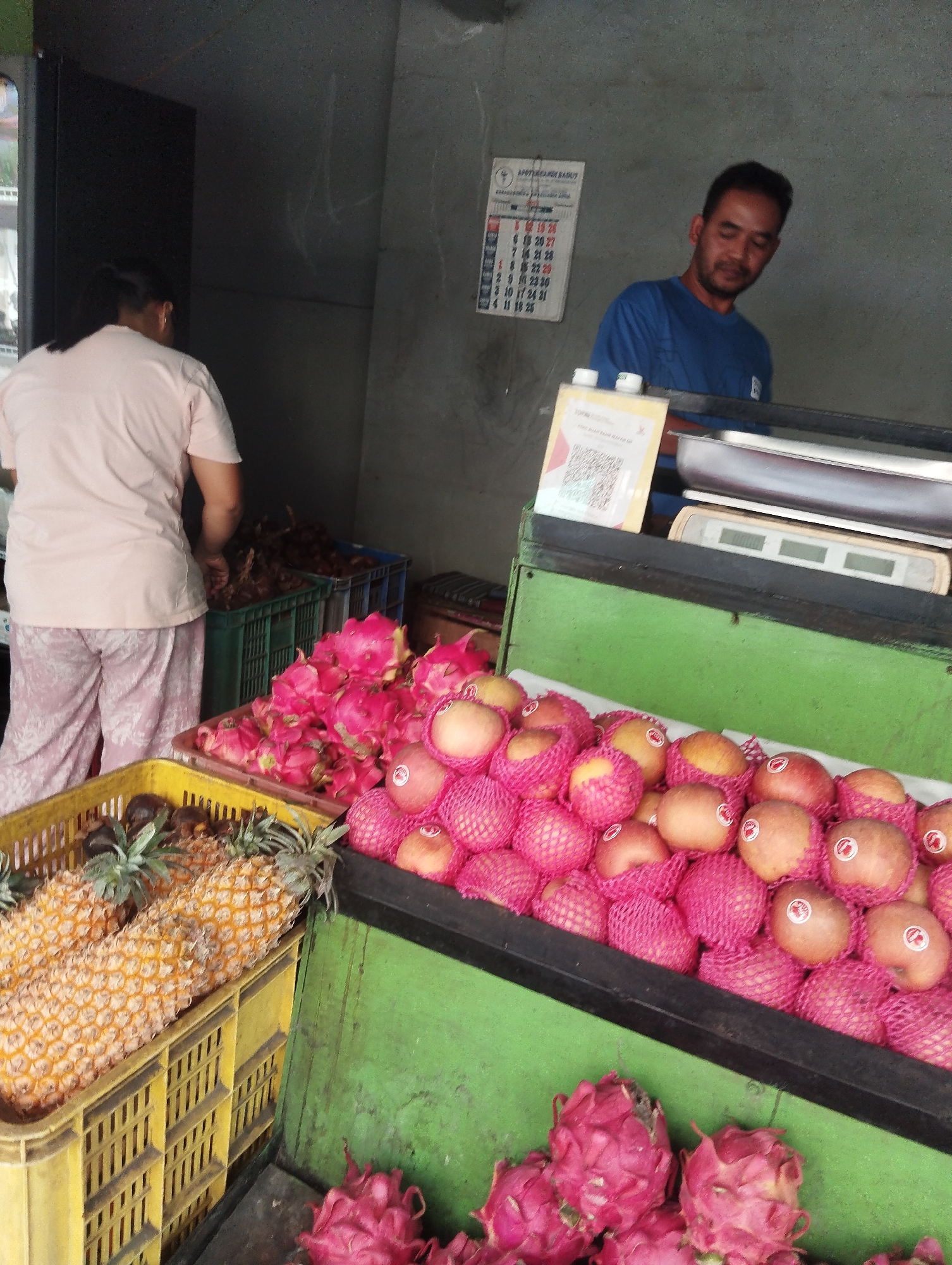 The image depicts a fruit stall at a market in Malang, Indonesia. In the foreground, several crates of fruit are visible. A yellow crate on the left contains two large pineapples. To the right of the pineapples, a black shelf displays numerous apples individually wrapped in pink netting. Below this shelf, a bright green surface holds a large pile of pink dragon fruit.
In the background, a woman wearing a light pink t-shirt and patterned pants is seen from the back, reaching into a dark green crate filled with what appears to be root vegetables or similar produce. Beside her, a man in a blue t-shirt stands behind a counter. On the wall behind the counter, a calendar is hung. A digital scale with a metallic tray is positioned on the counter, and a QR code stand is placed in front of it. The overall environment suggests an outdoor or semi-open market setting.