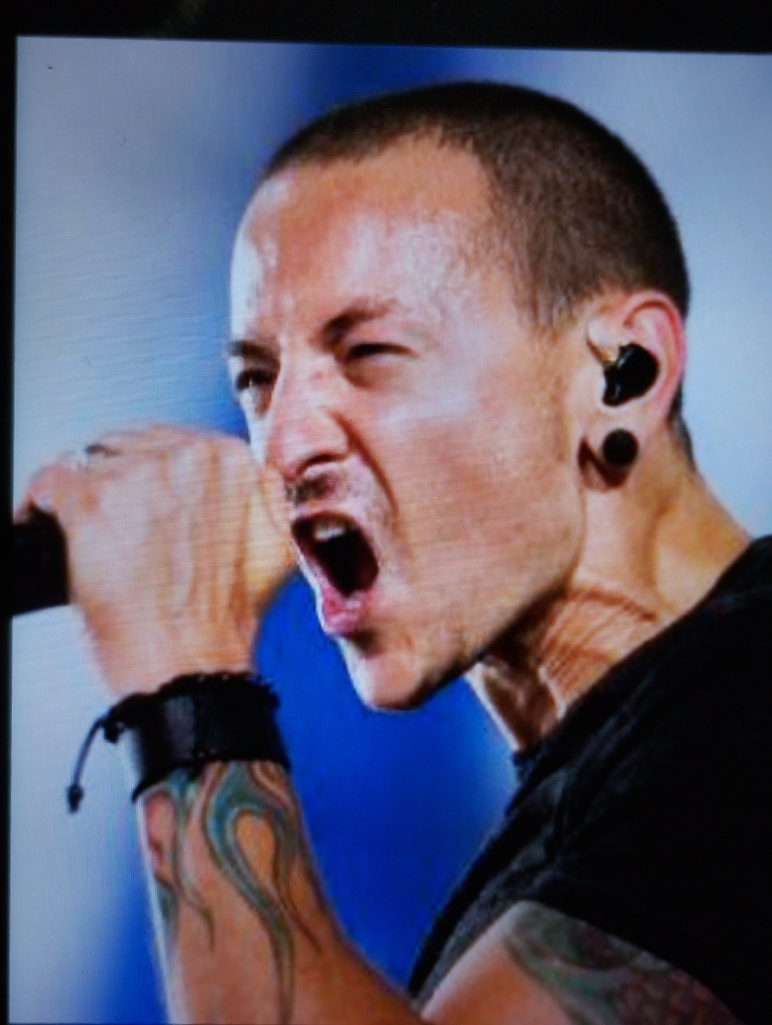 A close-up, head and shoulders, medium shot photograph captures a light-skinned male with short, dark hair and a prominent tattoo on his left forearm. He is wearing a black t-shirt and a black leather wristband. His mouth is open in a scream, revealing his teeth and tongue. He has a black earbud in his right ear and a stretched earlobe with a black plug. His left hand is raised and appears to be gripping a microphone, though the microphone itself is not fully visible. The background is a gradient of light blue to white. The image is slightly blurry and appears to have been taken from a screen.