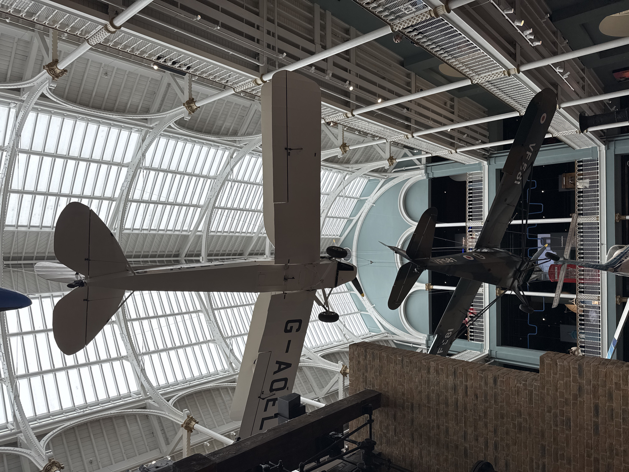 The image shows the interior of a museum, likely the National Museum of Scotland, given the architectural style and the presence of suspended aircraft. Two vintage aircraft are prominently displayed, suspended from the ceiling of a large atrium with a glass roof and ornate white metalwork. The upper aircraft is a biplane painted white with the registration 'G-ADFL' visible on its wing. Below it, another biplane, painted dark green and bearing the markings 'VF581' and what appears to be a Royal Air Force roundel, is also suspended. The atrium features multiple levels of balconies with white railings, overlooking the central space. People can be seen on the balconies. The overall impression is one of a grand exhibition hall designed to showcase historical artifacts. The architecture suggests a Victorian or Edwardian era building. The lighting is natural, filtered through the glass roof, and supplemented by artificial lights on the balconies.