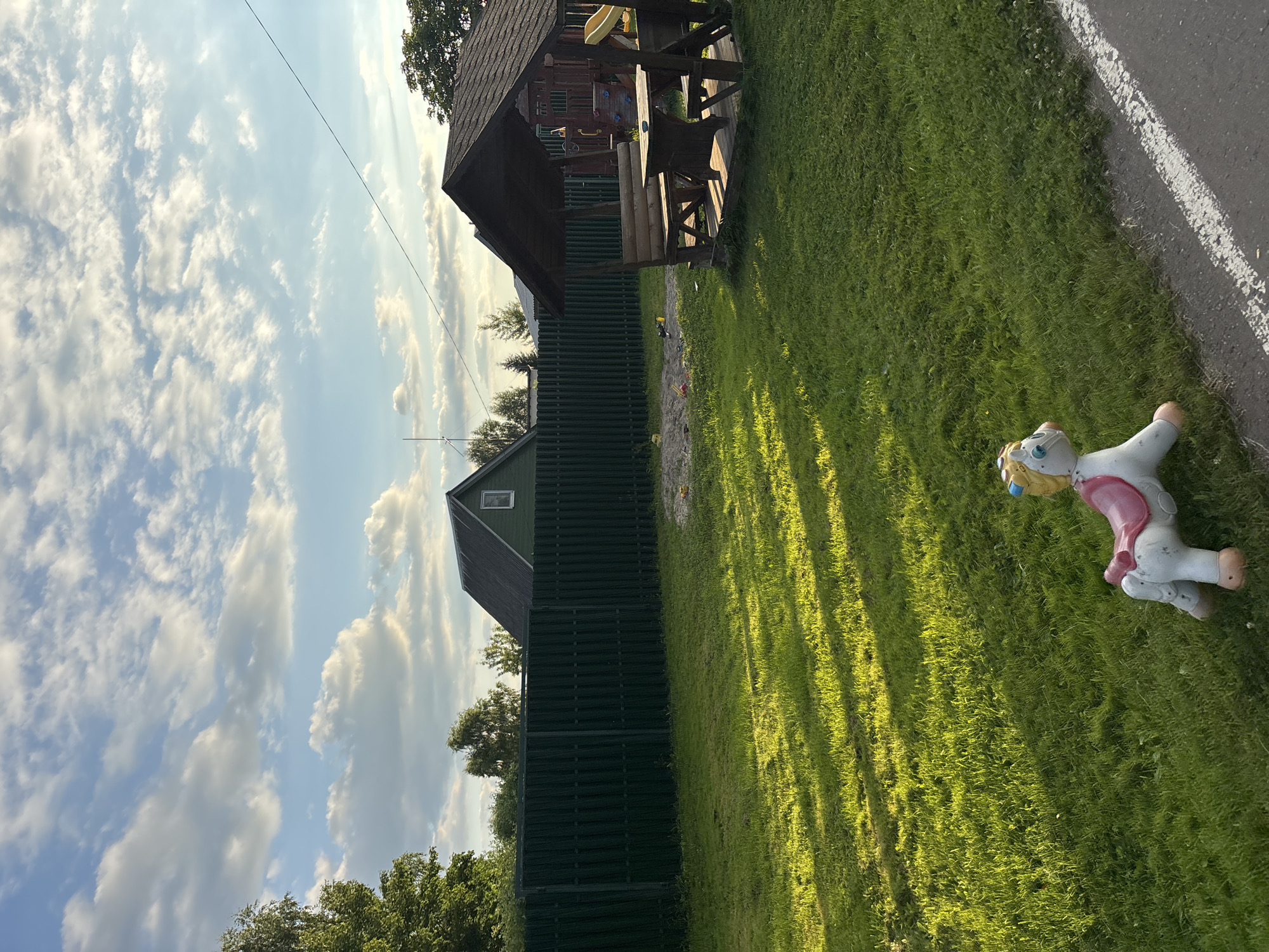 This media file captures a sunlit outdoor scene in Мещёры, Russia, likely during a warm day given the bright sunshine and lush green grass. The image is oriented vertically.
The primary subjects include a wooden playhouse with a dark roof, a picnic table under a covered area, and a green slatted fence. In the foreground, on the right side of the image, a white and pink toy horse is resting on the grass. The horse appears to be a children's toy, possibly a rocker or spring rider, with a yellow mane and blue eyes.
The setting is a grassy yard with trees visible in the background, suggesting a residential or recreational area. The sky is partly cloudy with fluffy white clouds scattered across a bright blue expanse. The sun is casting long shadows across the lawn, indicating it might be late afternoon or early morning. A paved surface with a white line marking is visible on the far right edge of the image. There is no visible text in the image. The overall mood is peaceful and summery.