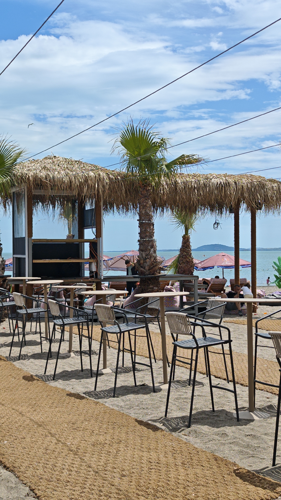 The image shows an outdoor beach bar or restaurant located in Burgas, Bulgaria. In the foreground, there are several high-top tables with bar stools arranged along a sandy area covered with a brown woven mat pathway. The tables are empty, suggesting the scene is either before or after peak hours. Behind the tables, there is a thatched-roof structure providing shade for a bar area. The roof is supported by wooden pillars. A palm tree grows through the roof, adding to the tropical ambiance.
In the background, the sea is visible with people relaxing on lounge chairs under parasols. The weather is sunny with a partly cloudy sky. A small island or land mass can be seen in the distance on the horizon. Power or communication lines are visible above the thatched roof. The scene appears relaxed and tourist-oriented, characteristic of a beachside establishment in Burgas.