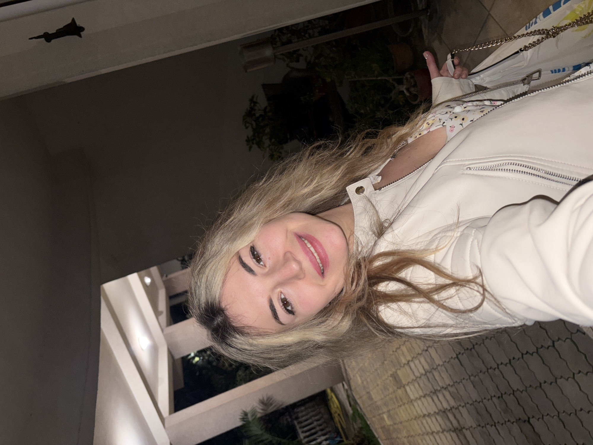 A young woman with blonde hair is smiling at the camera. She is wearing a white leather jacket over a floral-patterned top. A chain strap of a purse or bag is visible. The setting appears to be outdoors at night, with a walkway paved with interlocking stones and illuminated structures. The presence of plants and building architecture suggests a location in an urban environment. There is no clear evidence to establish a direct connection to Budva, Montenegro from this single image.