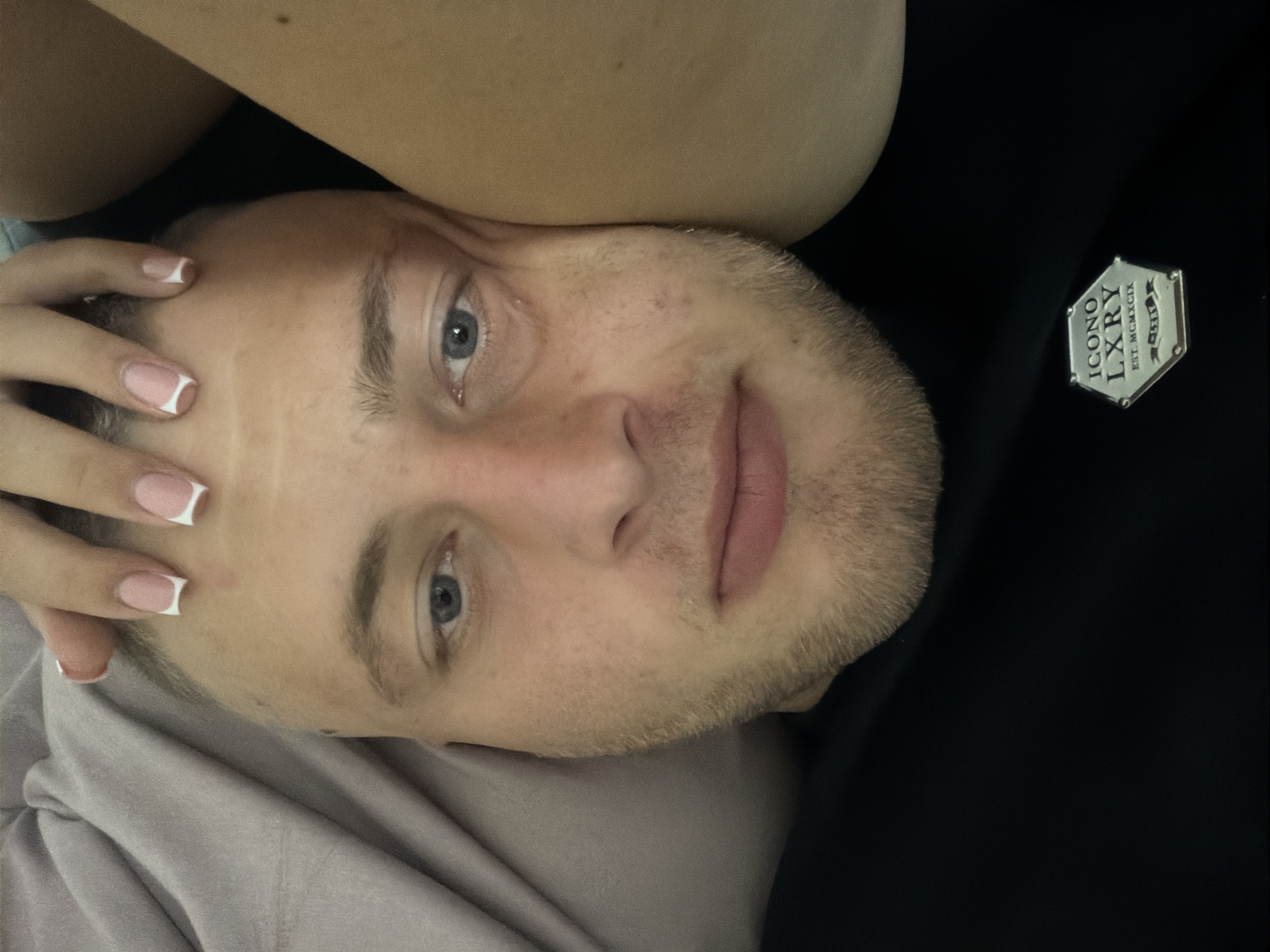 A close-up, eye-level shot shows a fair-skinned male lying down, his face turned towards the camera. His eyes are open and blue, and he has short, light brown hair. A faint stubble is visible on his chin and jawline. His lips are closed. A woman's hand with french manicured nails rests on his forehead. To the right of the man's face, a portion of a person's shoulder or upper arm is visible, providing a soft, blurred backdrop. The man is wearing a black t-shirt with a silver hexagonal emblem on the chest. The emblem has the words "ICONO LXRY" and "EST. MCMXCIX" inscribed on it. A light purple fabric, possibly a blanket or clothing, is visible on the left side of the frame, behind the man's head. The lighting is soft and diffused. No specific location in Strășeni, Moldova can be identified from the image.