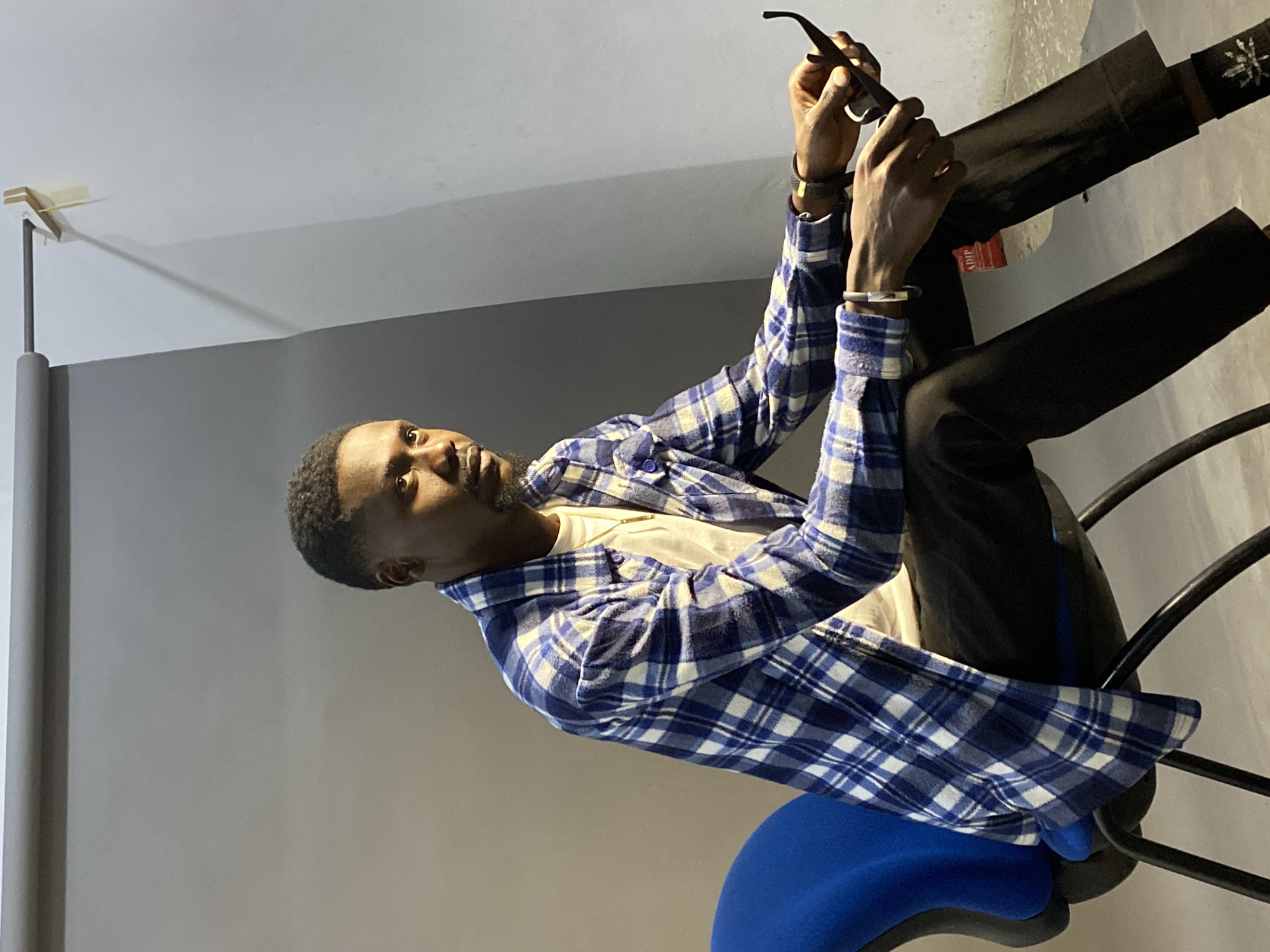 A man sits on a blue office chair. He is wearing black pants, a white t-shirt, and a blue and white plaid button-down shirt. He is holding a pair of sunglasses in his hands and looking at them. The background is a light gray wall with a darker gray section at the top. There is a lamp with a white shade on the left side of the image. The location appears to be indoors in a room with a modern interior. The city of Jos, Nigeria is not indicated.