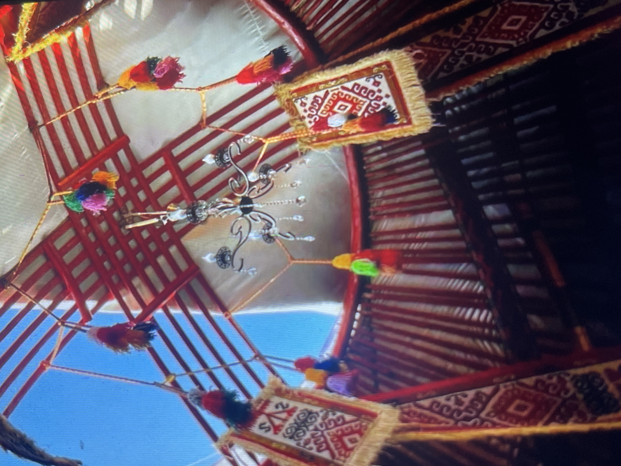 The image shows the interior of a yurt, a traditional nomadic dwelling. The ceiling structure, made of red wooden poles, is visible, as well as the white fabric covering. Several colorful tassels and small, decorative rugs hang from ropes attached to the ceiling. One rug on the left features a black and white geometric pattern with red accents, and another on the right has a similar design in red and white on a yellow background. A chandelier with crystal pendants hangs from the center of the ceiling. The yurt's interior walls are lined with vertical red and white striped fabric. The overall impression is of a well-decorated and possibly inhabited yurt. This scene is likely located in Bishkek City, Kyrgyzstan, given the context of yurts being a significant part of Kyrgyz culture. There are no people visible in the image.