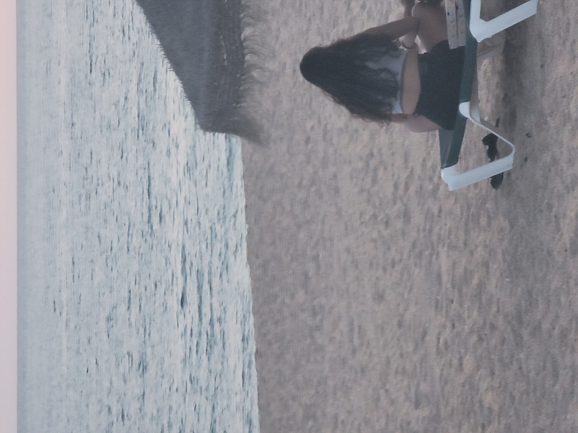 The image shows a woman sitting on a beach chair on a sandy beach in Quarteira, Portugal. The ocean is visible in the background with a gentle wave pattern and a pale sky. A thatched umbrella is partially visible on the right side of the image. The woman is facing away from the camera, with her dark hair down. She is wearing a light-colored crop top and dark shorts. Her legs are crossed as she sits on the green and white beach chair. The overall lighting suggests it may be early morning or late evening.