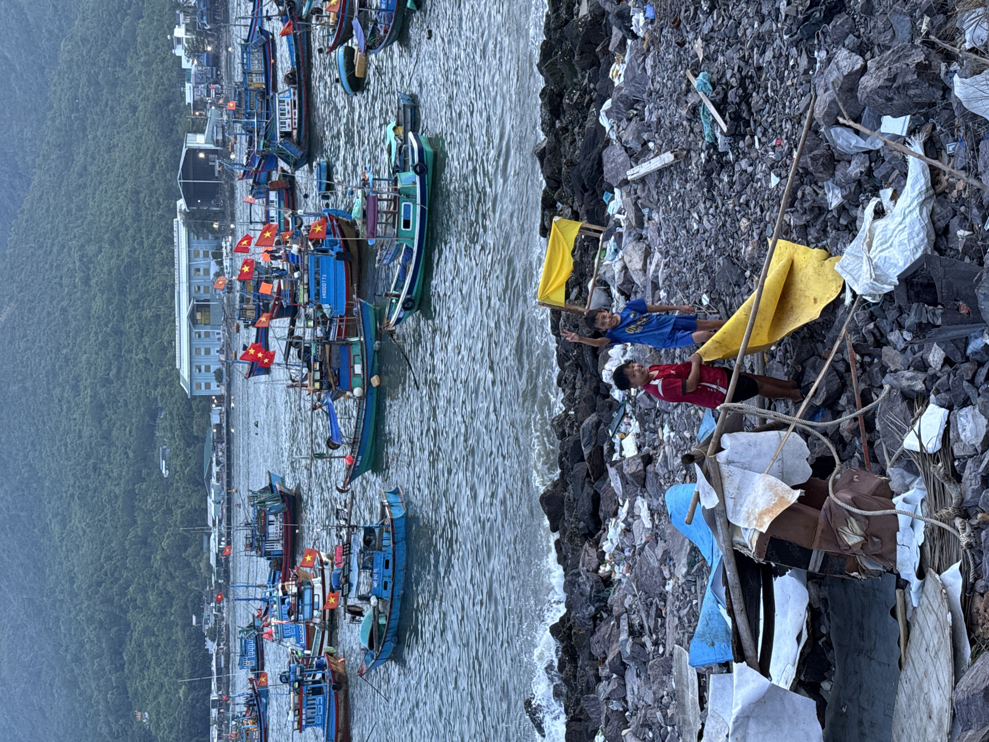 In Nha Trang, Vietnam, a coastline littered with debris from fishing operations and general refuse provides a stark foreground to a busy harbor filled with numerous fishing boats. Two young boys stand on the rocky shore; one in a blue shirt and shorts, the other in a red shirt and shorts. The boy in blue holds up a yellow flag and appears to be waving or making a peace sign, while the boy in red looks towards the viewer. The water is choppy, with waves breaking against the rocky shore, carrying with them plastic waste and other refuse. The boats, varying in size and color, are clustered together in the bay, many adorned with Vietnamese flags. In the background, a lush, green mountainside rises behind a line of buildings and industrial structures, suggesting a densely populated coastal area. The overall scene captures the daily life and economic activity of a Vietnamese fishing community, juxtaposed with the environmental challenges posed by pollution.