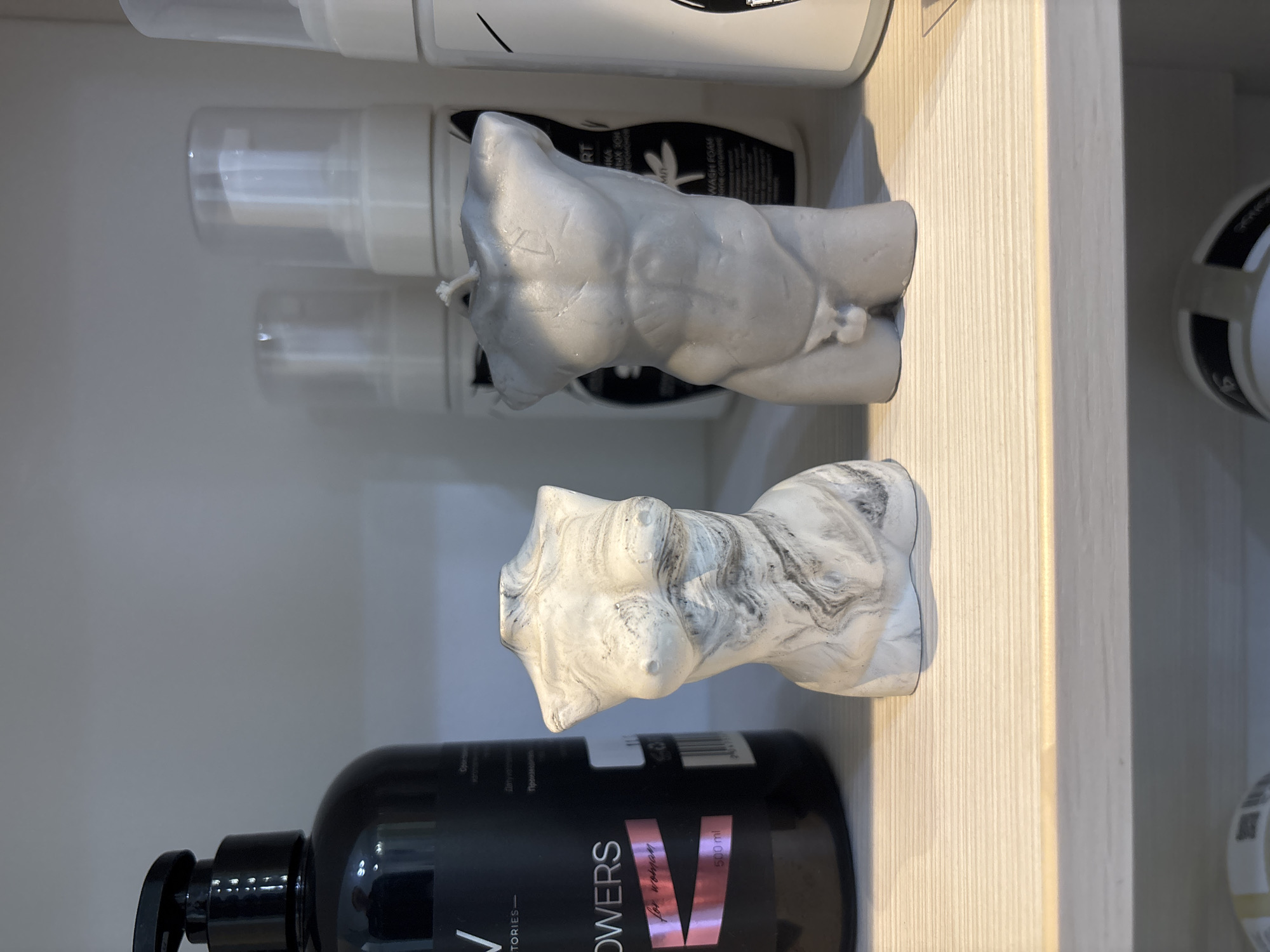 The image shows a shelf with various items. There are bottles of beauty products, specifically foam wash, arranged vertically on the shelf. Two sculptural objects resembling torsos, one in a gray color and the other with a marbled white and gray pattern, are positioned in front of the bottles. The lighting suggests the scene may be indoors, and the items appear to be neatly organized. The environment appears to be a clean, well-lit shelf, likely in a retail or personal storage space. It is not possible to locate it in Vsevolozhsk, Russia, with only this picture.