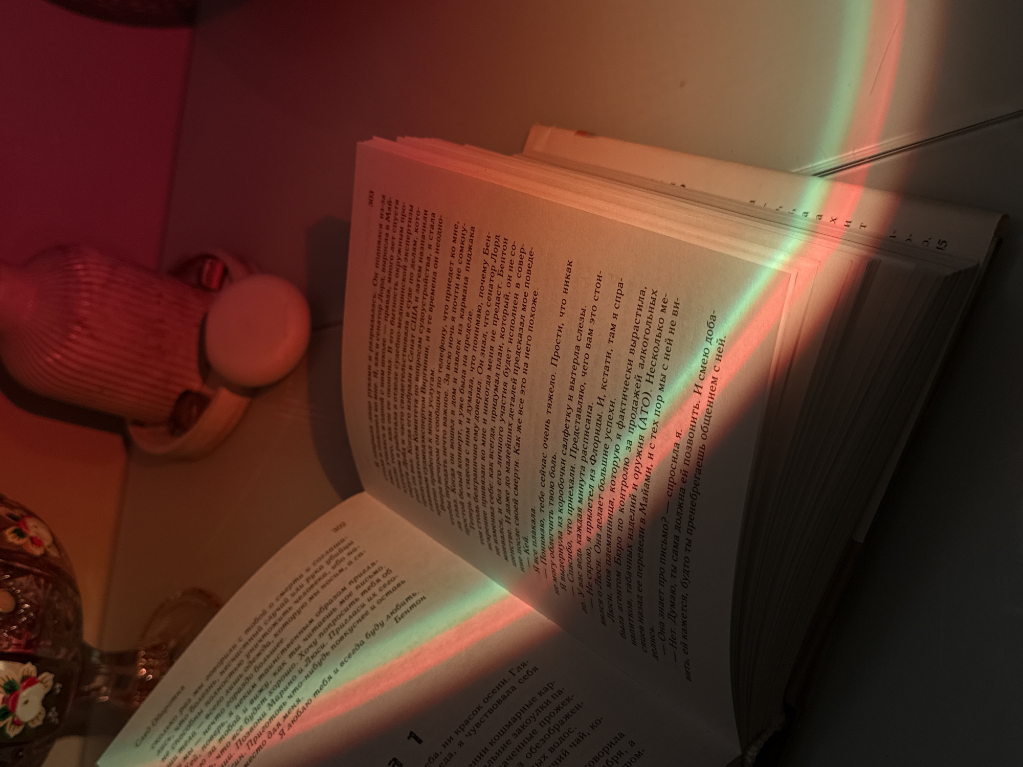 An open book, likely a novel written in Russian, lies on a surface, illuminated by striking rainbow-colored light. The light, featuring distinct bands of red, orange, and green, casts vibrant hues across the white pages of the book, creating an ethereal glow. The book is open to pages 302 and 303.
On the left page (302), text titled "След Оборотня" (Trace of the Werewolf/Werewolf's Trail) is visible. The content appears to be a personal letter or reflection, discussing themes of death, illness, and human existence, concluding with the sign-off "Я люблю тебя и всегда буду любить. Бентон" (I love you and will always love you. Benton). The start of a new section or chapter is visible at the bottom of this page, mentioning "неба, ни красок осени" (sky, nor colors of autumn).
The right page (303) continues the narrative, mentioning crying, a "Senator Lord," and the city of Miami. It refers to a "Bureau for the control of alcoholic beverages, tobacco products and weapons (ATO)" and dialogue about a letter: "Она знает про письмо?" (Does she know about the letter?) and "Нет. Думаю, ты сама должна ей позвонить." (No. I think you should call her yourself.). A character named "Lucy" (Люси), described as a niece, is also mentioned.
Other objects in the dimly lit scene include a ribbed, reddish-orange vase or bottle positioned behind the book on the left. Next to the vase is a small, light-colored, oval object. In the bottom left corner, part of a decorative object, possibly ceramic or glass with floral patterns, is visible. The overall ambiance is quiet and contemplative, with the rainbow light adding a unique, almost magical, touch to the indoor setting. The time of day is not explicitly clear, but the atmospheric lighting suggests either a sunset, a prism catching light, or specific artificial illumination. Location context is unknown.