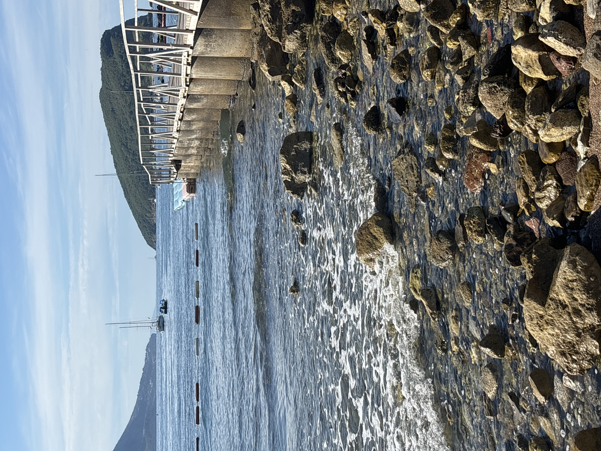This is a photograph of a rocky shoreline with a pier extending into the calm blue water. The pier, made of wood and supported by concrete pillars, has a white railing. In the distance, two sailboats are visible on the water. A small, wooded island with a gently sloping hillside is seen to the right of the pier, and a larger, mountainous landscape forms the backdrop to the left. The sky is a clear blue with a few wispy clouds. The water is clear enough to see the rocks beneath the surface, and small waves are gently lapping at the shore. A line of buoys, marking a swimming area, stretches across the water in the mid-ground. This scene is likely located on the coast of Budva, Montenegro, a popular tourist destination known for its scenic beaches and clear waters. There are no people visible in the image.