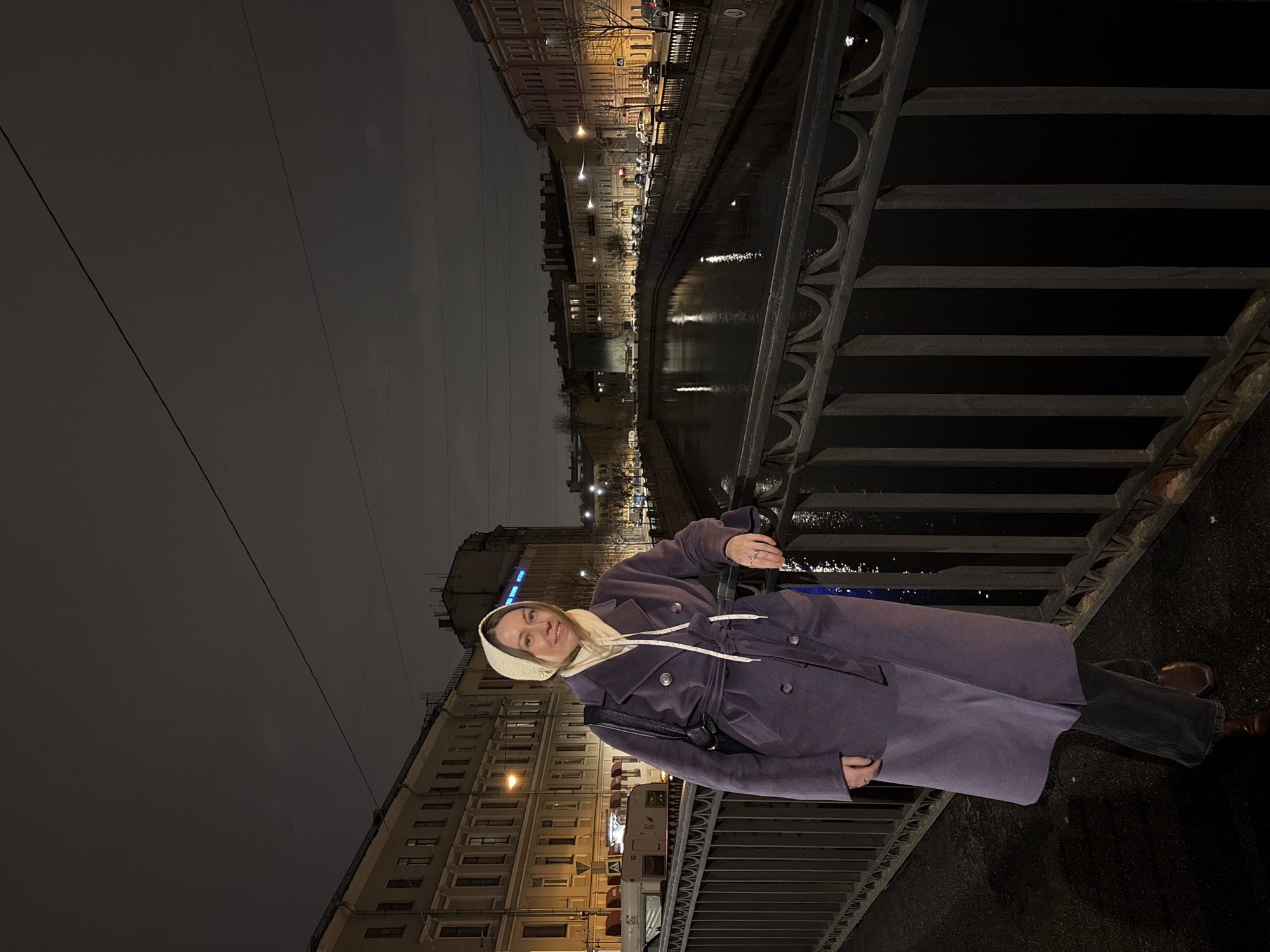 A woman stands on a bridge in Saint Petersburg, Russia. She wears a long purple coat and a light-colored headscarf. The bridge has a dark metal railing. Below, a canal reflects the city lights, which illuminate buildings that line the waterway. The sky is dark and overcast.