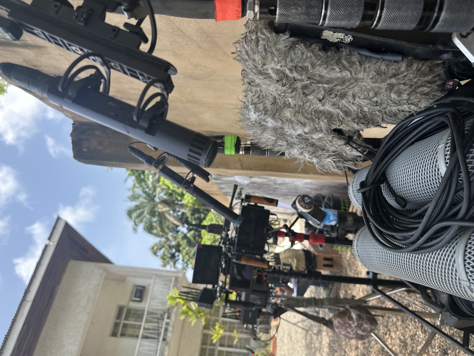 The image shows a filming set-up outdoors with audio equipment prominently displayed, including shotgun microphones with windscreens and coiled cables. The equipment is mounted on stands. In the background, a white building with windows is visible, along with green trees and a blue sky with clouds. The location appears to be Lagos, Nigeria.