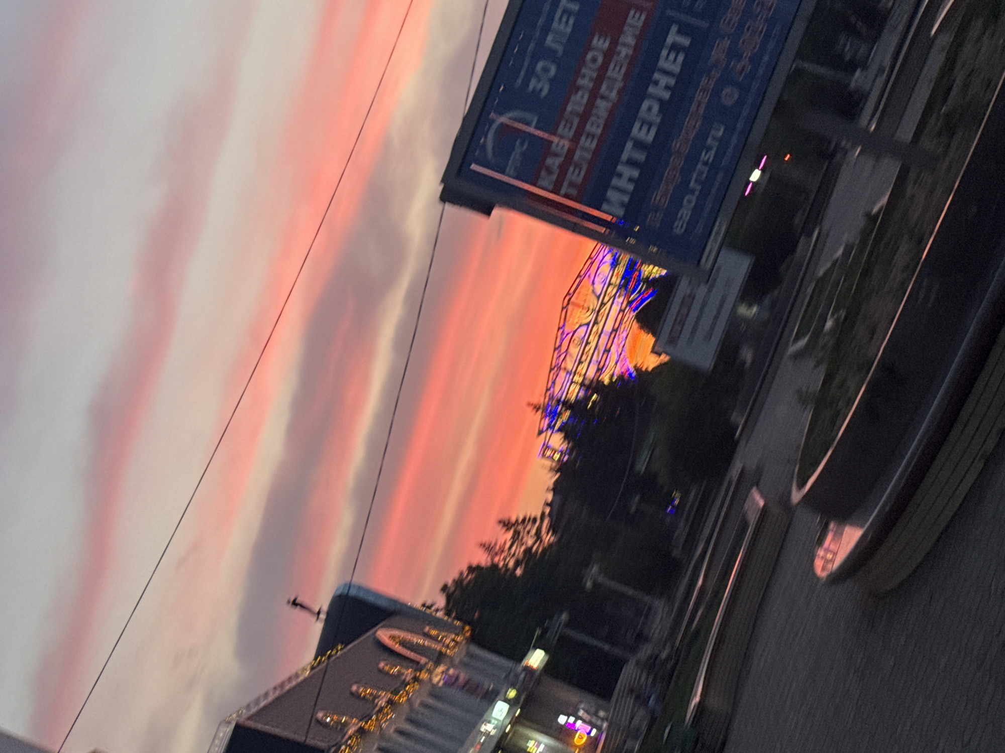 The image captures a street scene in Birobidzhan, Russia, during a sunset. The sky is filled with vibrant pink and orange hues, indicative of a colorful twilight. In the foreground, a paved area with planters is visible. To the left, a building with illuminated decorative lights and arched windows suggests a commercial or public establishment. Behind it, trees are silhouetted against the sky. To the right, a blue billboard with text in Russian, including "ИНТЕРНЕТ" (INTERNET), is prominently displayed. A Ferris wheel-like structure, illuminated with colorful lights, is visible in the middle ground. Overhead power lines traverse the sky. The overall impression is an urban environment at dusk with elements of infrastructure and recreation.