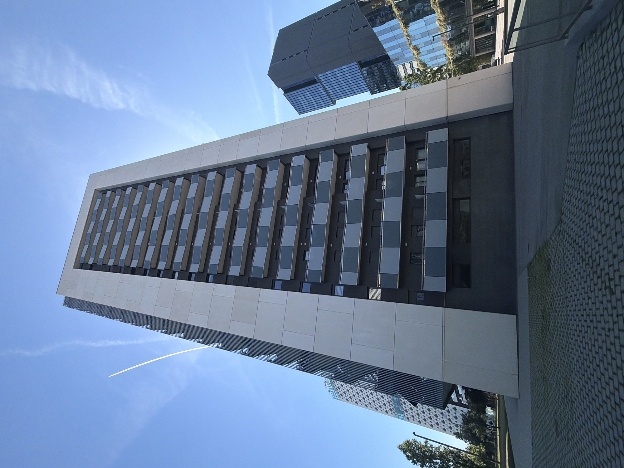 The image shows a modern building in l'Hospitalet de Llobregat, Spain. The building has a rectangular shape, with a light-colored frame and darker, patterned panels in between. There are rows of alternating light and dark panels, creating a checkerboard effect. Another building, partially obscured, is reflected in the windows of the closer building. The sky is blue with faint contrails from an airplane. A section of cobblestone pavement is visible in the lower right of the frame. Trees and parts of other buildings are visible in the background.