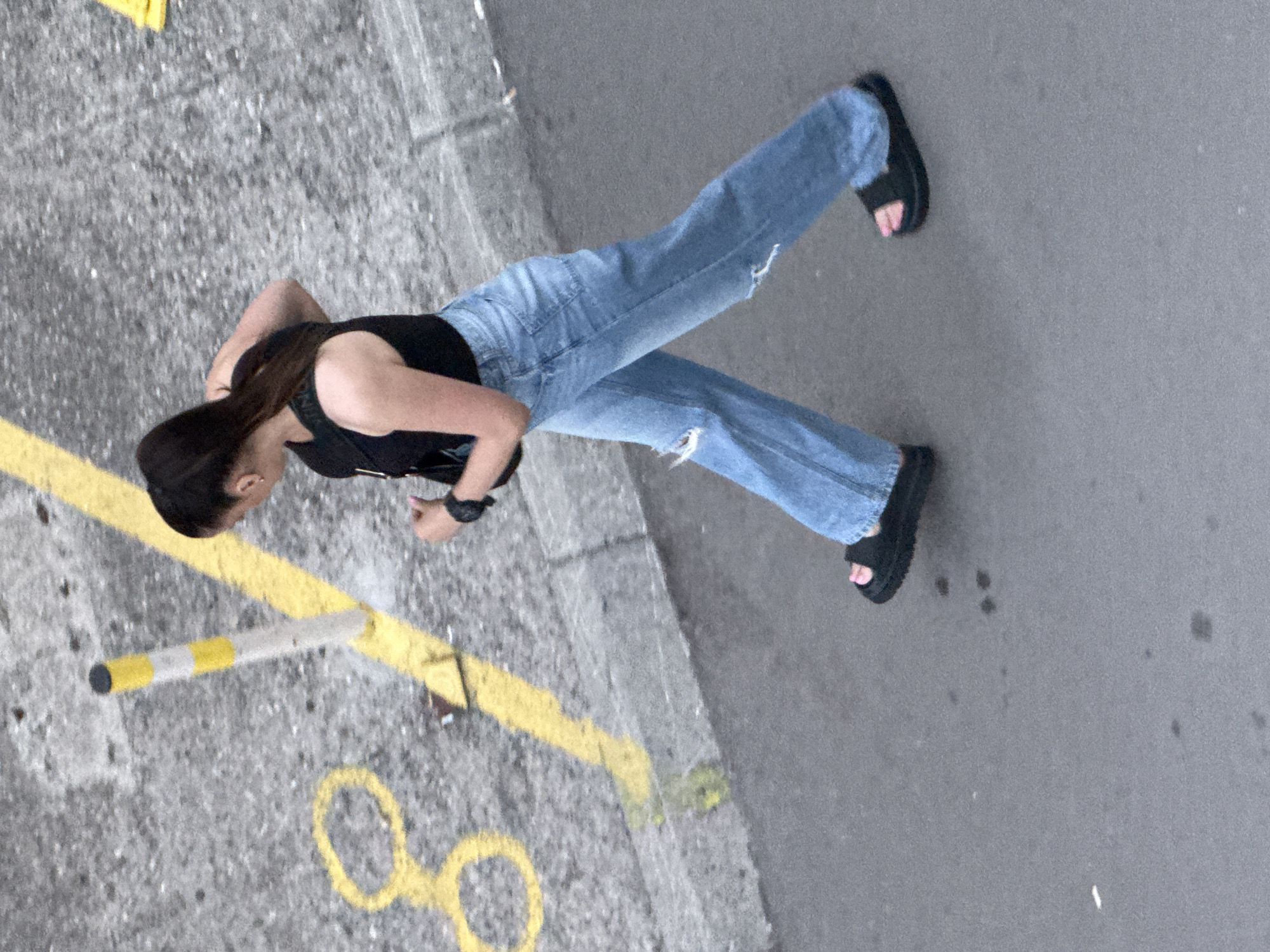An outdoor, high-angle, medium shot captures a young woman walking on a paved surface in Budva, Montenegro. The woman, with her dark hair pulled back in a ponytail, is wearing a black tank top, light blue distressed jeans, and black slide sandals. She is carrying a black crossbody bag. The ground is a rough, gray asphalt with yellow painted markings, including a broken circle and a diagonal line. A yellow and white striped bollard is also visible on the left side of the frame. The scene appears to be a street or walkway with concrete curbing.