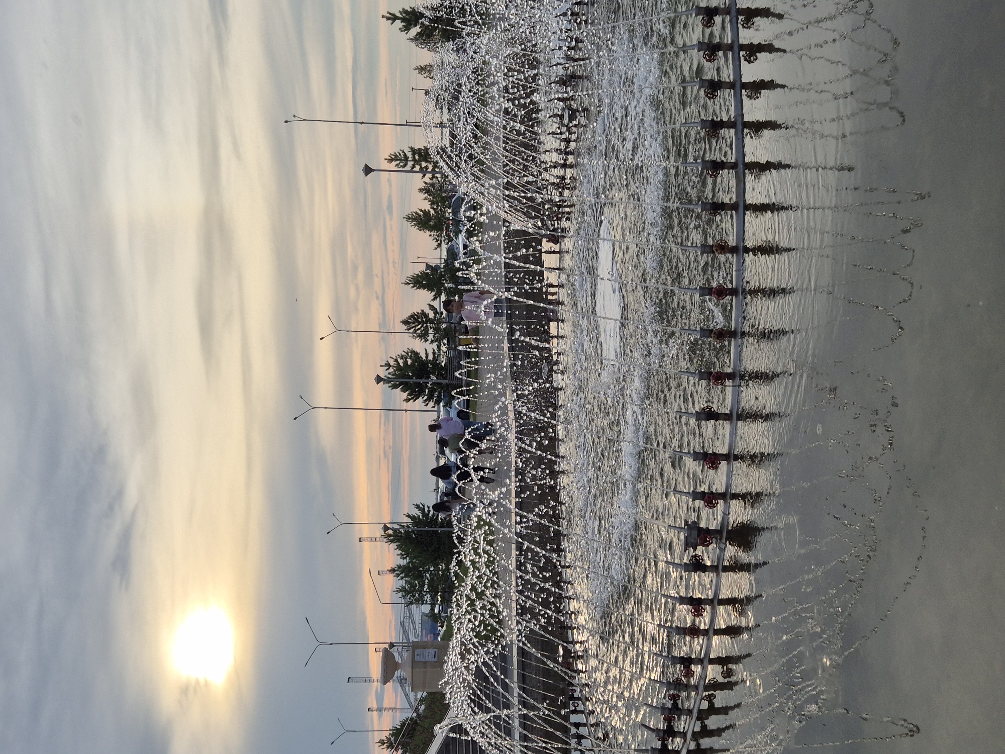 The image depicts a street scene in Kokshetau, Kazakhstan, at sunset. The foreground is dominated by a large fountain with numerous jets of water creating a shimmering, curtain-like effect. The water spray reflects the warm hues of the setting sun. Behind the fountain, a paved walkway is visible with several people strolling or standing. A row of trees lines the walkway, beyond which more trees and some buildings can be seen. Streetlights are spaced along the walkway. The sky is filled with dramatic clouds, tinged with orange and yellow from the setting sun, which is a bright, glowing orb on the left side of the frame. The overall mood is serene and picturesque, capturing the beauty of an evening in an urban park. No visible text is present in the image.