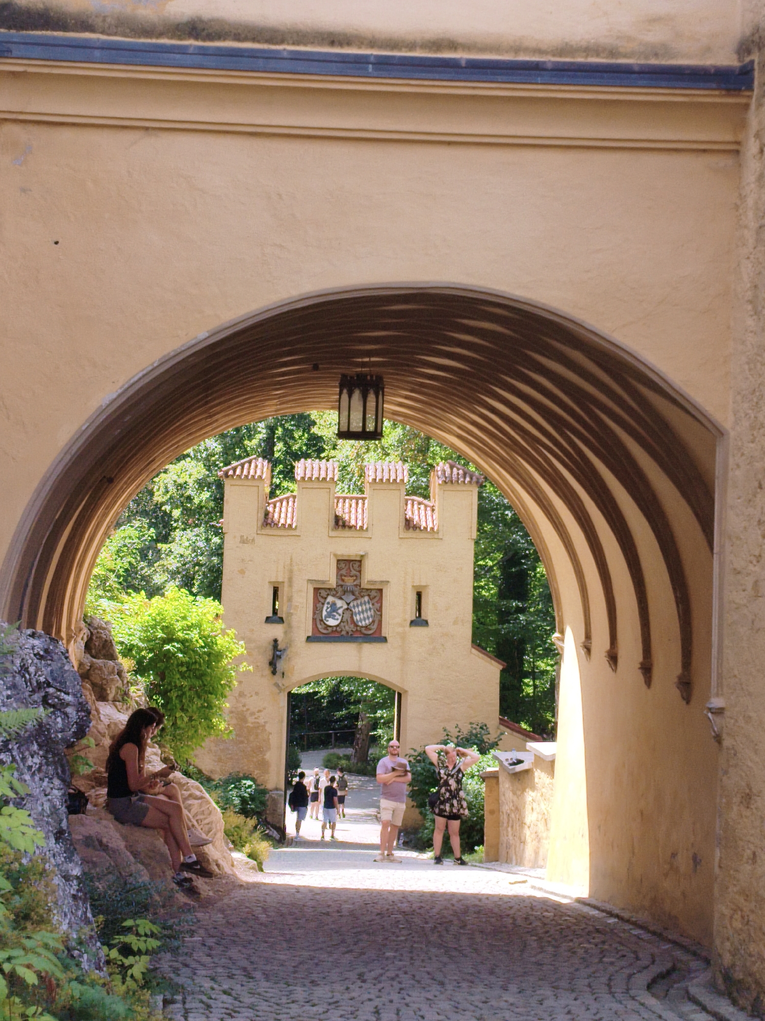 This is a vertical, eye-level, full-color photograph of an arched passageway leading towards a yellow castle in Schwangau, Germany. The passageway is constructed of tan-colored stone with a ribbed, arched ceiling. A dark, lantern-style light fixture hangs from the ceiling of the arch.
In the foreground, a woman with long brown hair is sitting on a rock on the left side of the path, wearing a black tank top and gray shorts. She is holding an object in her hands. The path is paved with cobblestones.
In the middle ground, through the arch, a section of the castle is visible. It is yellow with red-tiled roofs and features a crest with two shield emblems above an arched entryway. Several people are walking through the passageway, including a man in a purple t-shirt and shorts on the right, and a woman in a floral dress with her hands behind her head. Behind them, several other people are walking in the distance.
The background is filled with lush green trees. The lighting suggests it is daytime with sunlight casting shadows. The overall impression is a scenic view of a historic location.