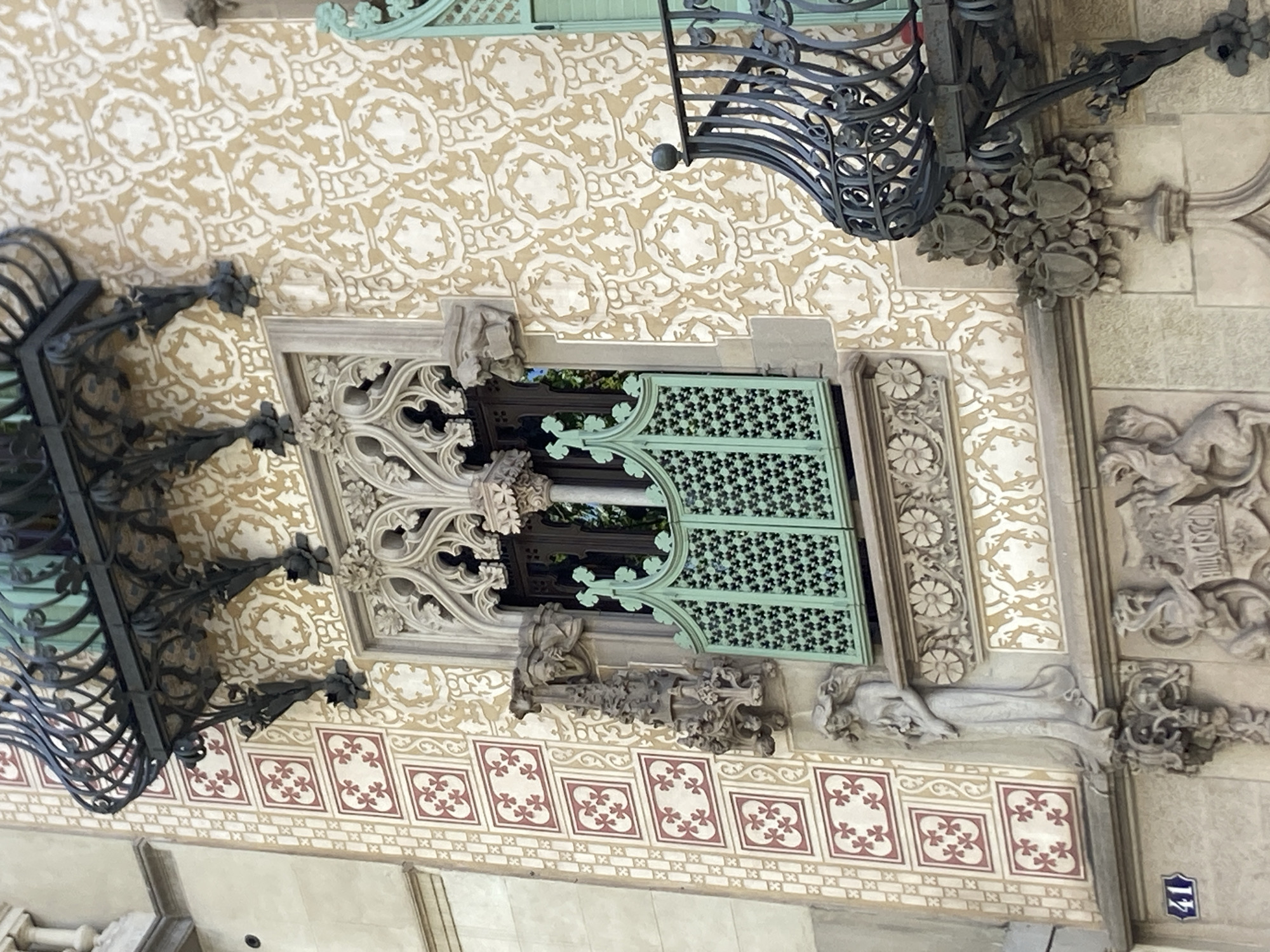 The image shows an ornate architectural detail on the facade of a building in Barcelona, Spain. The focus is on a Gothic-style window with decorative stonework and a bright green metal grille. The surrounding wall is adorned with a repeating cream-colored pattern on a beige background, and a section to the left features a tilework pattern of red crosses within squares. Above the window is a wrought-iron balcony with intricate scrollwork. Below the window, a carved stone figure of a woman holding a bouquet stands on a decorative corbel. Further down, the building's lower facade is visible, with more sculpted elements and a street number "41" displayed on a blue and white tile. The overall impression is one of rich, detailed historical architecture.