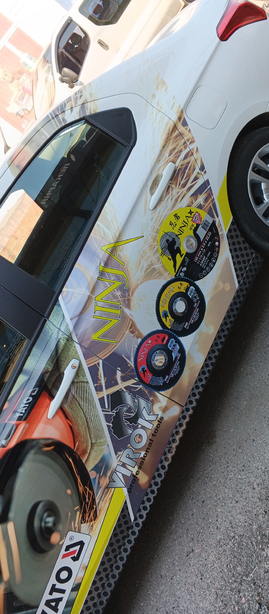The image depicts a white car with a graphic wrap on its side. The wrap features imagery related to metalworking, including sparks, a grinder, and cutting discs. The word "NINJA" is prominently displayed in large yellow lettering across the side of the car. Several circular cutting discs, branded "NINJA" and other manufacturers, are affixed to the car door. The car appears to be parked outdoors, with a grey, textured surface, possibly asphalt or concrete, visible beneath it. The image is taken from a slightly elevated angle, looking down at the car. The location is identified as Smila, Ukraine. There are no people clearly visible in the image, and the focus is on the car and its graphic design.