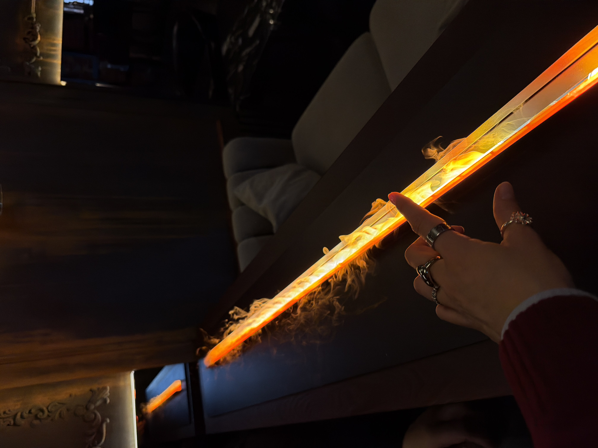 The image shows a dark, interior scene. In the foreground, a hand with multiple rings is reaching towards a long, rectangular light source that casts a warm orange glow, resembling a light bar or strip. Some smoke appears to be emanating from the light source. The background contains a blurred interior with what seems to be upholstered furniture and dark walls. The location is Moscow, Russia, as it is identified by the AI analysis as such.