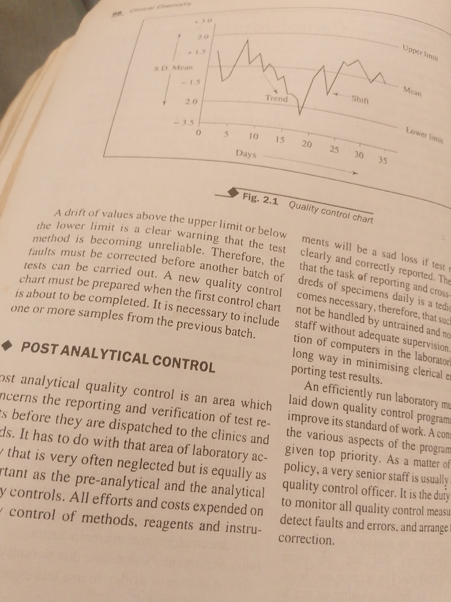 The image shows an open book with text and a graph. The graph is a quality control chart, labeled "Fig. 2.1 Quality control chart", depicting "Days" on the x-axis and values with "S.D. Mean" on the y-axis, with lines indicating "Upper limit", "Mean", and "Lower limit". The graph also labels "Trend" and "Shift". The text discusses laboratory procedures, quality control, and the importance of accurate reporting and error detection. The text appears to be from a textbook on Clinical Chemistry, as indicated by the heading "Clinical Chemistry". The setting is likely a study or office environment where someone is reading or working with this book. There is no indication of people, specific time of day, weather, or emotions. The location context provided is Jalingo, Nigeria, but there are no visual cues in the image to confirm this specific location.