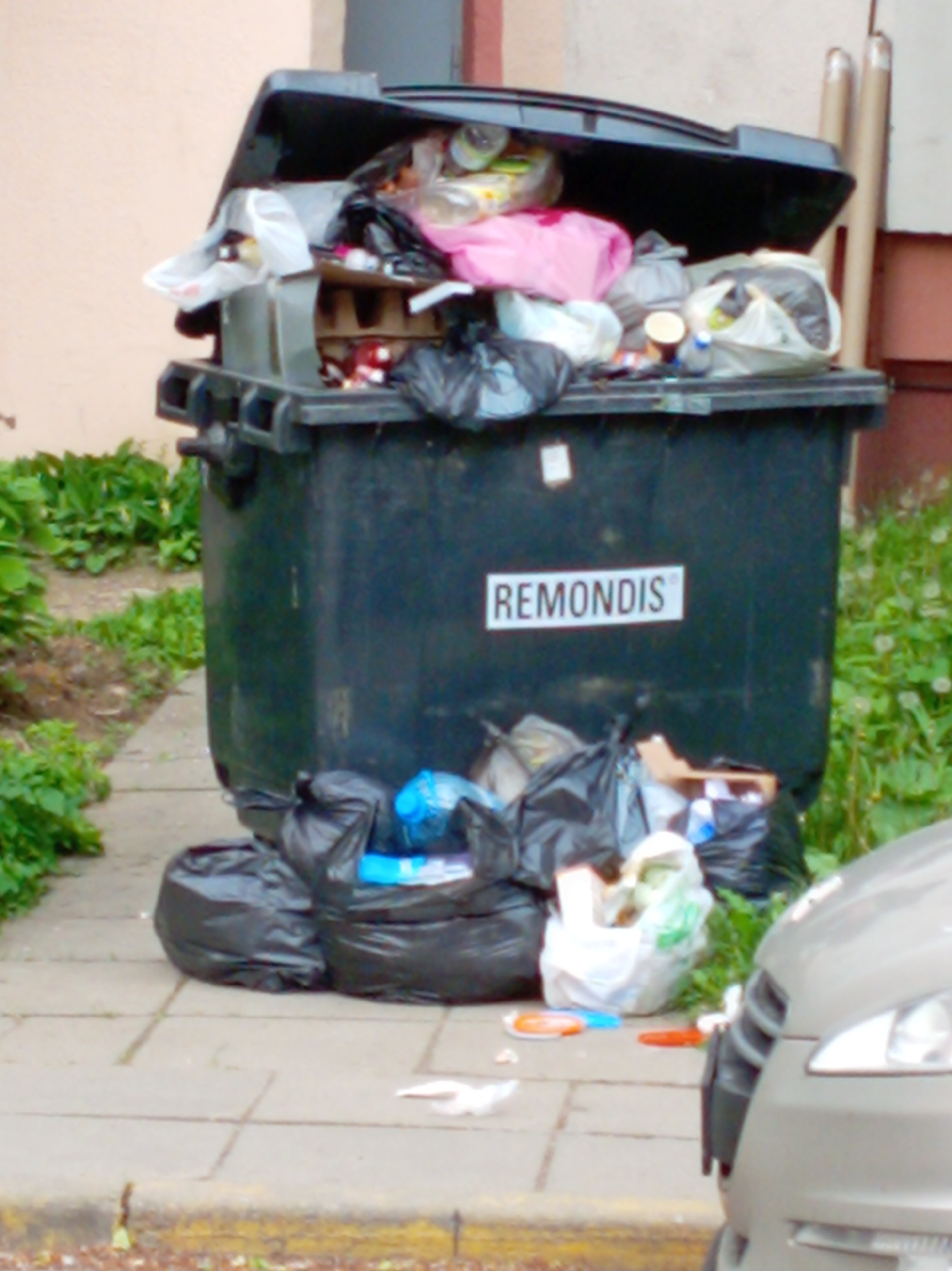 The image depicts an overflowing black trash bin, labeled "REMONDIS", standing on a paved surface in what appears to be an urban environment. The lid of the bin is partially open, revealing a variety of waste, including plastic bags, cardboard, and food containers. Several black plastic trash bags are clustered at the base of the bin on the paved surface. Scattered around the base are pieces of litter, including discarded plastic items and paper. A portion of a car is visible to the right side of the image. The background shows a building with a peach-colored exterior and greenery. The location appears to be Unknown, Unknown.