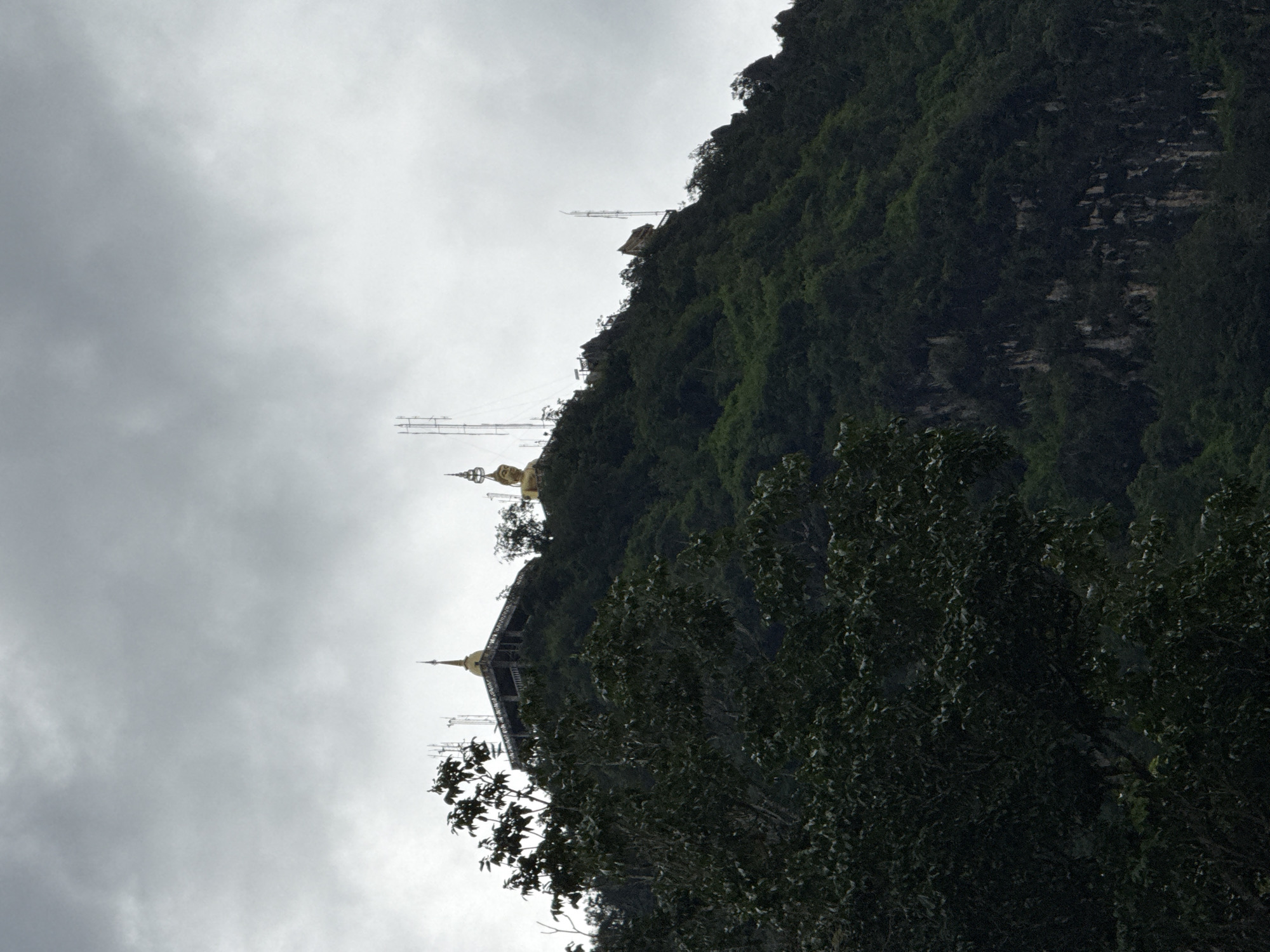 This is a wide shot of a lush, green mountain in Ban Khao Tang, Thailand, under a heavily overcast sky. The mountain is covered in dense foliage, with sheer rock faces visible in places. At the summit, there is a temple complex with a golden pagoda, and a large golden statue of the Buddha. Several communication towers are also present on the mountain. In the foreground, the dark, leafy branches of a tree partially obscure the view of the mountain. The overall impression is one of a serene, natural landscape with religious and technological elements integrated into its rugged terrain.