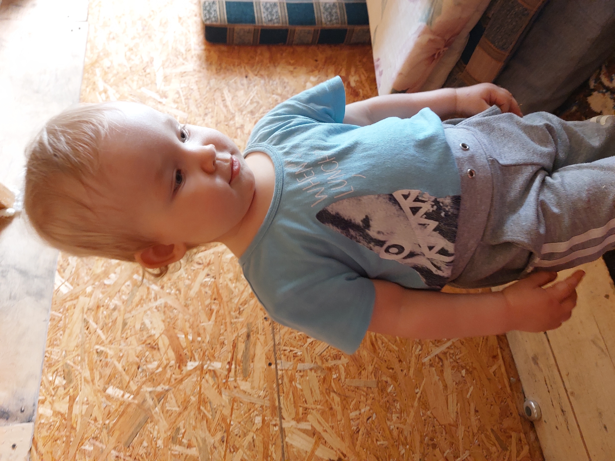 The image shows a young child lying on a floor made of oriented strand board (OSB). The child has blonde hair and is wearing a light blue t-shirt with a graphic print that includes text. The text on the t-shirt appears to say "WHEN LUNCH". The child is also wearing grey sweatpants.
In the background, there are upholstered cushions or a sofa, suggesting an indoor setting. The lighting appears to be natural, possibly from a window. The overall scene is informal and domestic.
The child's expression is relaxed, and they seem to be looking off to the side. There are no specific indicators of the time of day or weather conditions, other than the presence of natural light. The location cues are limited to the OSB flooring and the general indoor furniture. The child's pose and expression suggest a moment of rest or play.