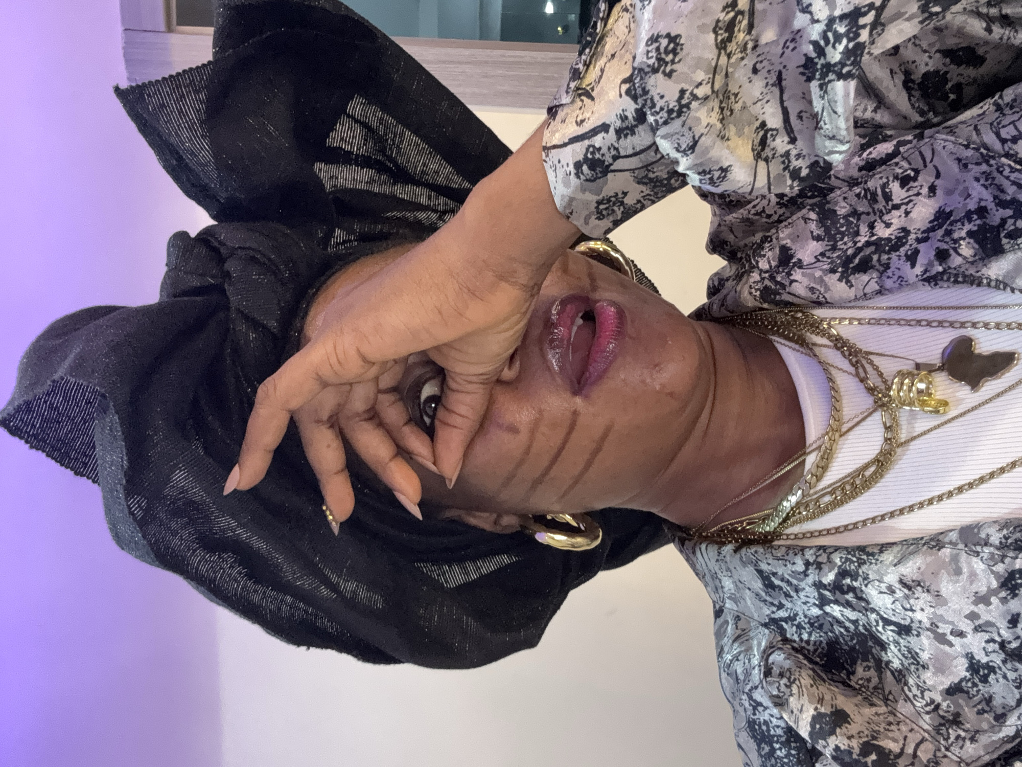A woman is pictured in a close-up shot. She is wearing a black headscarf, a patterned jacket, and multiple gold necklaces. Her right hand is raised, covering her right eye. She has three dark lines painted on her cheek. The background appears to be an indoor setting with a wall and a hint of a window or doorway. The lighting suggests artificial indoor lighting with a purple hue in the upper portion of the image. The image is focused on the woman's face and upper body. The city of Ajara, Nigeria is not depicted in this image.
