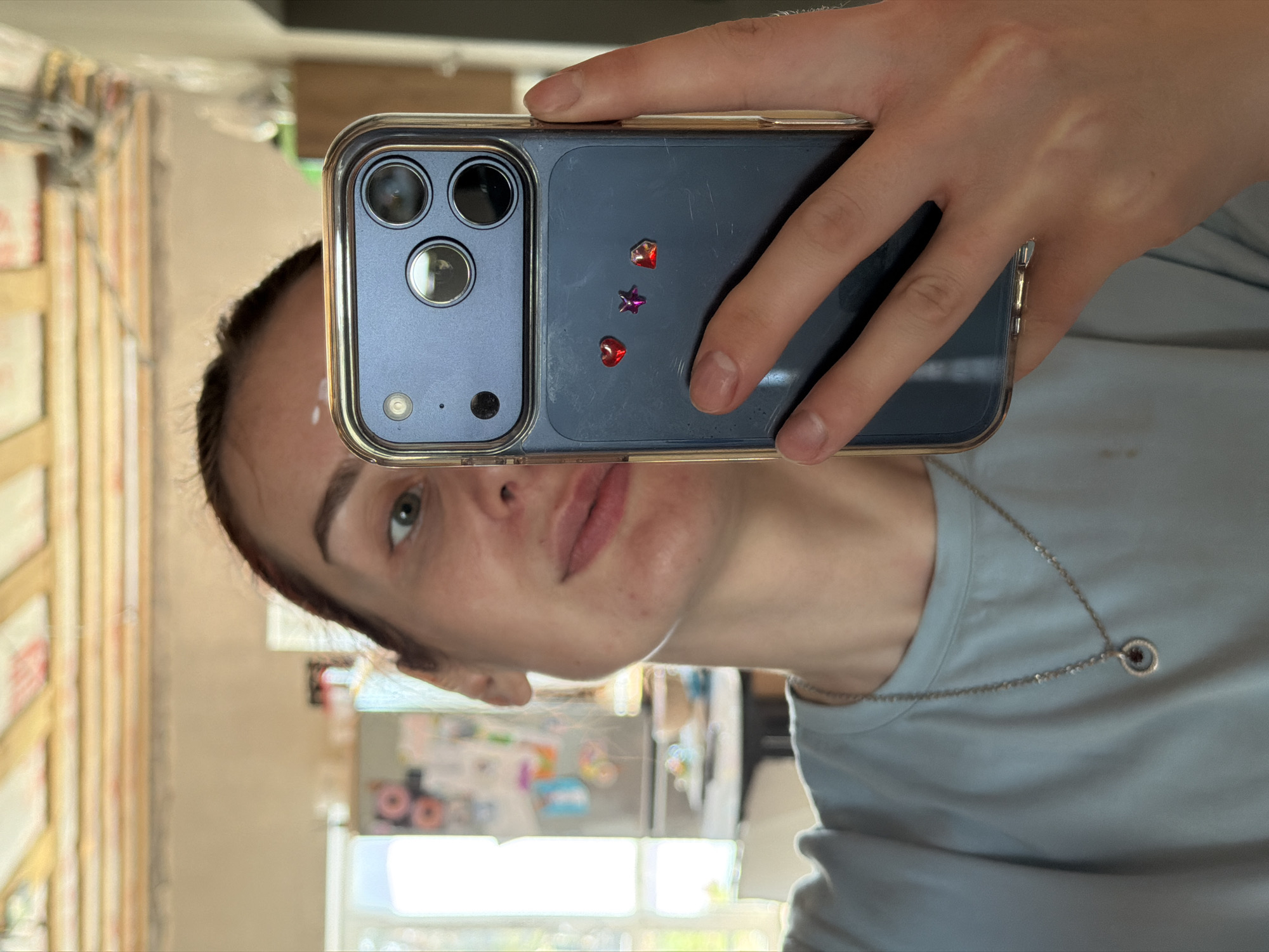 A person holds a smartphone in front of their face, taking a selfie. The phone is blue with a gold-colored case and decorated with a small red heart, a purple star, and a second red heart adhered to the back. The person has fair skin, light eyes, and dark hair pulled back from their face. They are wearing a light blue t-shirt and a silver necklace with a circular pendant. The background is a blurred interior scene, potentially a kitchen or dining area. The image is oriented sideways. There is a visible wall with wooden planks or paneling. The location is unspecified, but it's requested to be described as being in Chaikovsky, Russia.