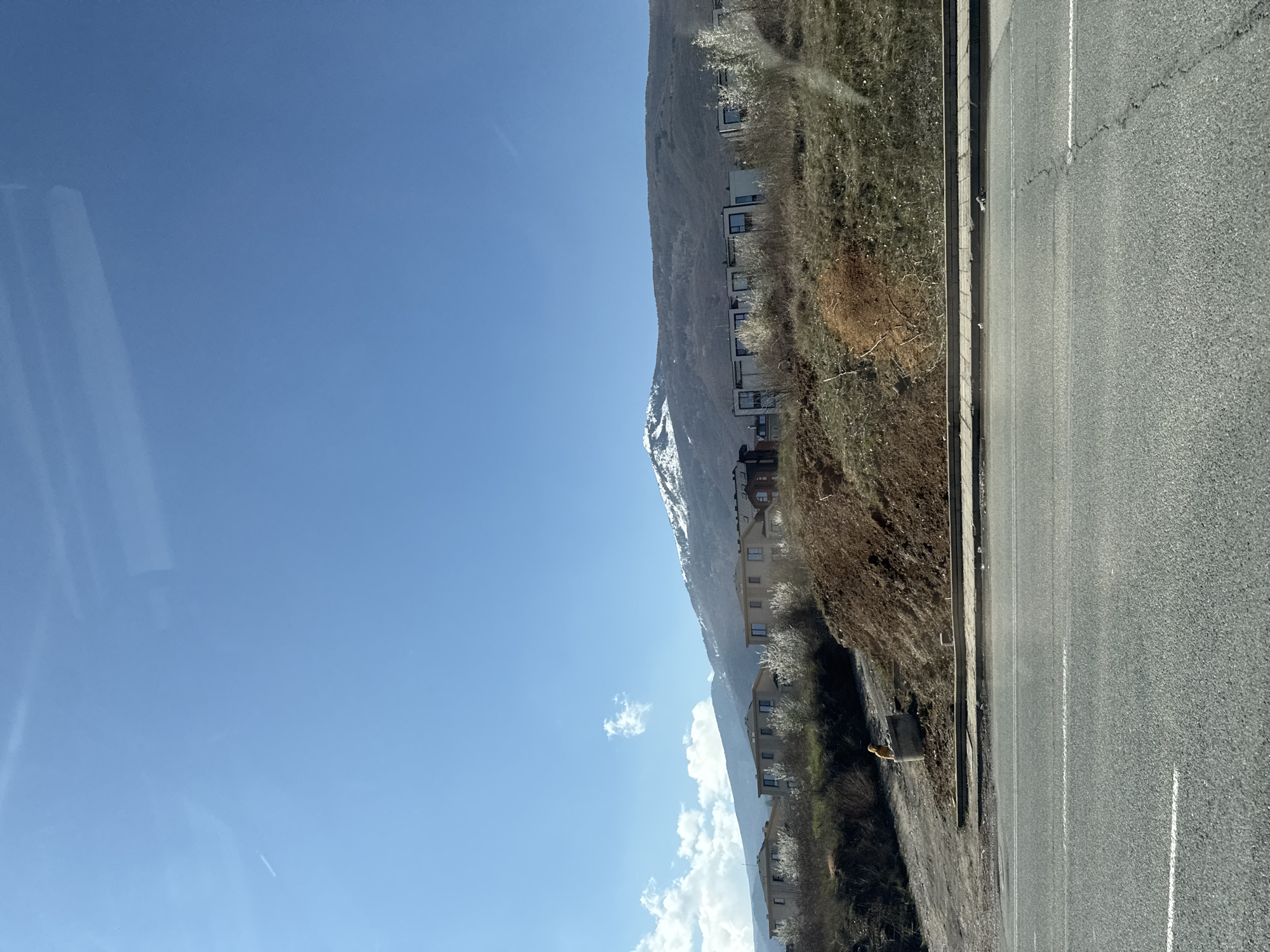 The image presents a landscape featuring a mountain range partially covered in snow, with residential buildings constructed along its lower slopes. A road runs adjacent to the hillside. The sky is clear with a few scattered clouds. It's difficult to pinpoint the precise location in Sofia, Bulgaria without additional context or features specific to the area. Based on the environment, this seems to be outside of the city center.