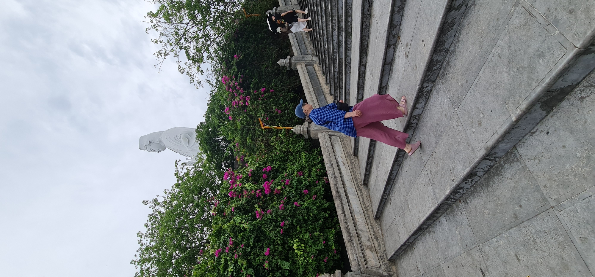 This outdoor image, taken in Đà Nẵng, Vietnam, captures a serene scene at a religious or tourist site. The central focus is a wide stone staircase ascending from the lower right towards the upper left. On the left side of the frame, partially obscured by lush green trees and vibrant pink bougainvillea flowers, stands a majestic, large white statue. The statue, likely depicting a Buddhist deity such as Guanyin, appears tranquil, with its head slightly bowed, facing towards the right.
On the stone staircase, a woman in the foreground is visible, dressed in a blue and white checkered shirt, loose-fitting maroon or pink pants, and matching pink sandals. She wears a blue baseball cap and carries a dark bag. She appears to be walking up the steps. Further up the staircase, two other individuals can be seen; one is holding a black umbrella and wearing a dark top with a white skirt, while the other is mostly hidden.
The sky overhead is overcast, filled with soft, diffuse clouds, suggesting either an early morning or late afternoon setting, or simply cloudy weather. The stone steps appear somewhat weathered and possibly damp, indicating recent rain or high humidity. The overall atmosphere of the scene is peaceful, combining natural beauty with architectural and spiritual elements. No visible text is present in the image.
