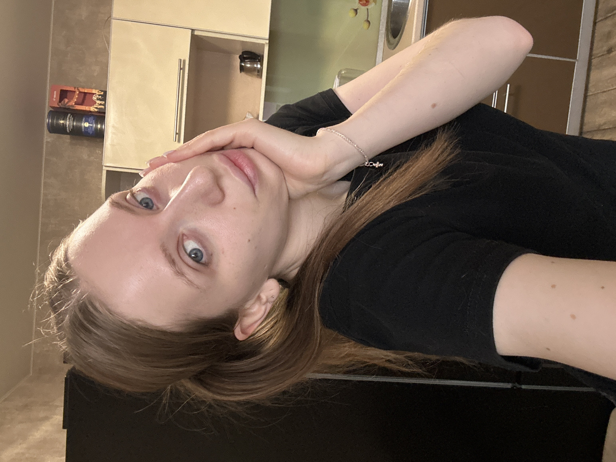 A woman with light skin and light brown hair is indoors, possibly in a kitchen. She wears a black shirt and a silver bracelet. Her face is close to the camera, her right hand is touching her face. Kitchen cabinets are visible in the background. There is no verifiable information to determine the location as Chaikovsky, Russia.