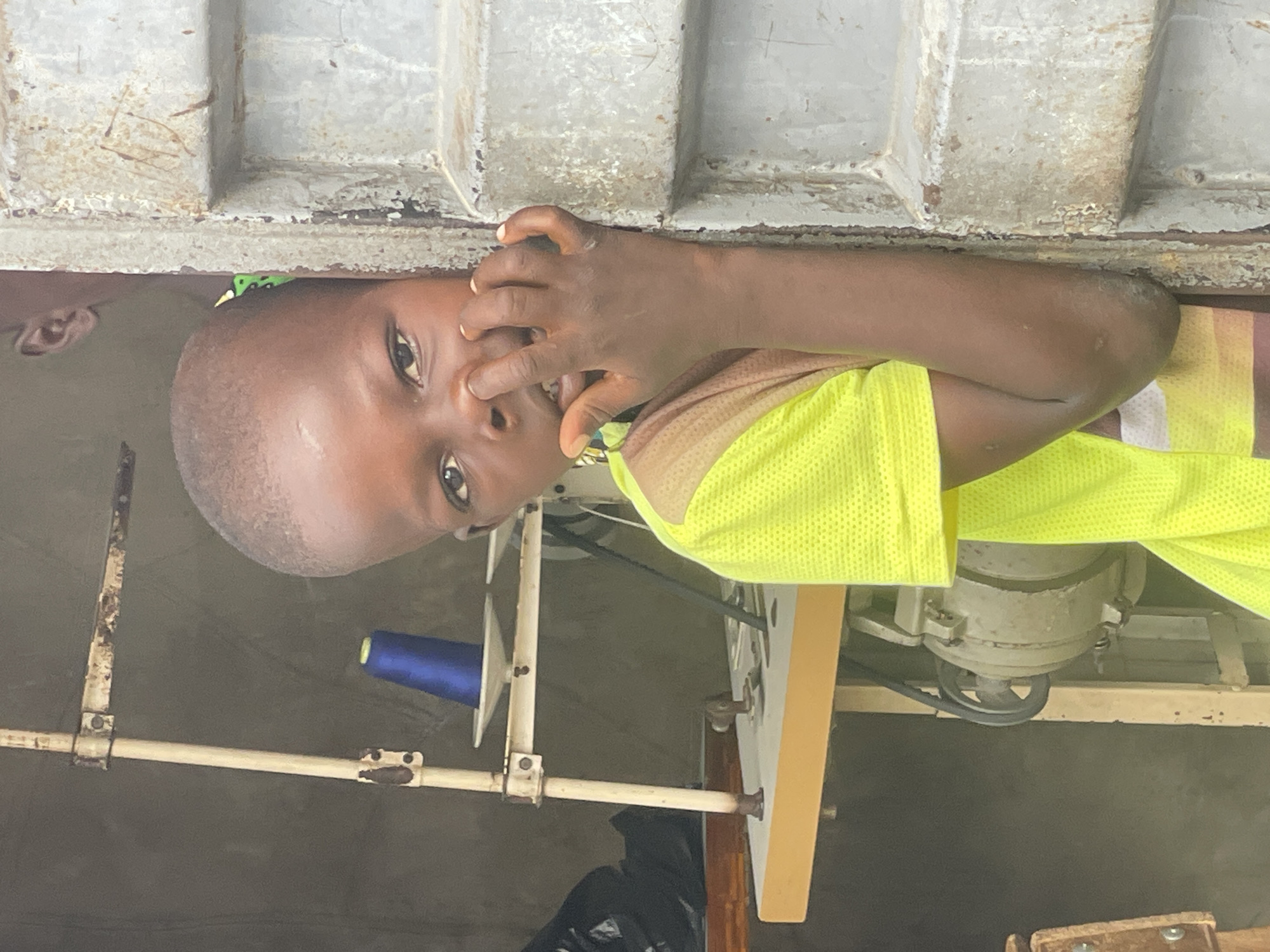 The image, rotated 90 degrees counter-clockwise, presents a close-up, waist-up portrait of a young child in what appears to be a workshop or market stall, likely in Ado, Nigeria.
The child, with dark skin and short, closely cropped hair, looks directly at the viewer with an expression of slight shyness or curiosity. Their right hand is raised, partially covering their mouth and nose, with fingers slightly splayed. They are wearing a vibrant, possibly neon, yellow mesh-like short-sleeved top over a lighter, possibly tan, inner layer. A small section of green fabric with a black pattern is visible on the top of their head, possibly part of a head covering or clothing detail. The ear of another person is faintly visible in the blurred background to the far left of the child.
The setting appears to be a functional workspace. To the right of the child (when viewed upright) is a sewing machine, partially visible, including its motor, a drive belt, and a light-colored wooden table or platform. A bright blue spool of thread is mounted on a metal spindle to the left of the child. Numerous metal structures, including rusty poles, bars, and frames, form part of the sewing machine stand or the general environment. A black plastic bag is visible beneath the sewing machine table. The background is dominated by a weathered, corrugated metal surface, showing significant rust and peeling paint, suggesting it is a wall or ceiling section of a basic structure.
The child is not engaged in a specific activity related to the sewing machine, but rather seems to be pausing to look at the camera. The overall scene conveys a candid moment in a working environment consistent with a small-scale tailoring or textile business in a community setting.