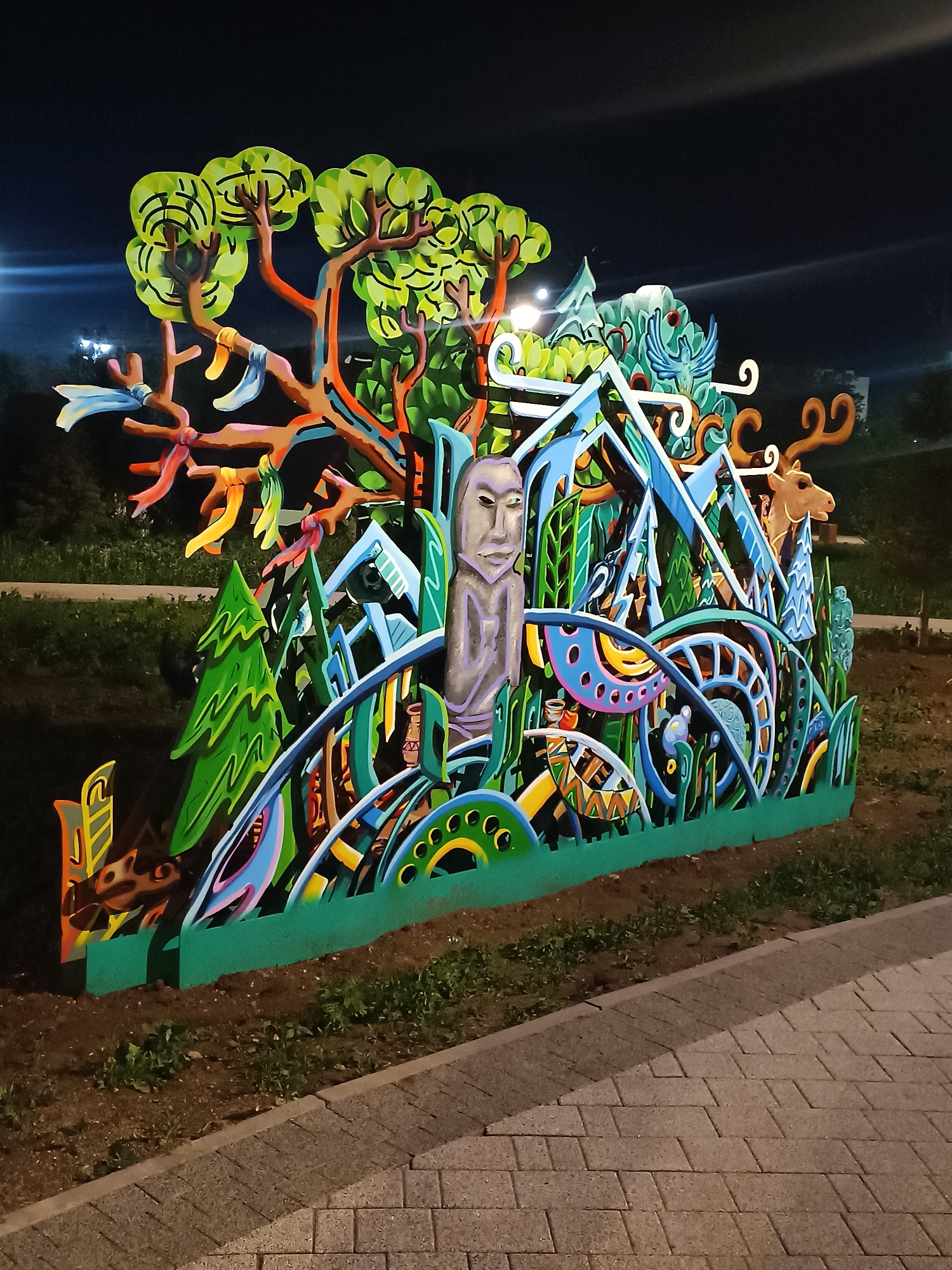 The image shows an outdoor art installation located in Kokshetau, Kazakhstan. The structure is a large, colorful, multi-layered piece of artwork depicting a landscape scene. Elements include trees with ribbons tied to the branches, abstract representations of mountains, stylized evergreens, and a human figure. The overall design is somewhat cartoonish and features various geometric patterns and bright colors. The installation appears to be made of painted wood or similar material. It is positioned on a grassy area adjacent to a paved walkway. The background is dark, suggesting the photo was taken at night. Streetlights are visible in the distance, illuminating the installation.