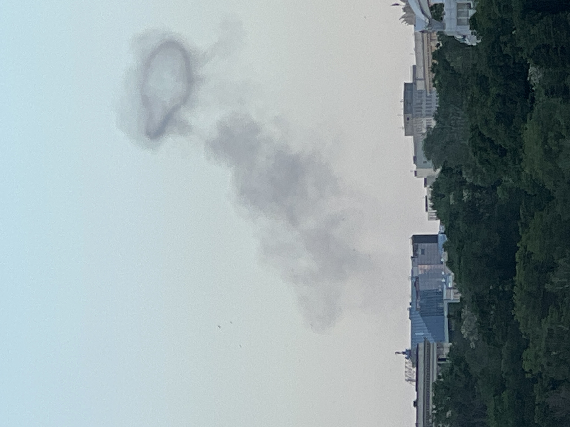 The image depicts a column of gray smoke in the sky above an urban environment. The smoke is denser towards the top, forming a distinct ring-like shape. Below the ring, the smoke thins out and diffuses downwards. In the background, buildings and trees are visible. The city's name is Unknown, Unknown.