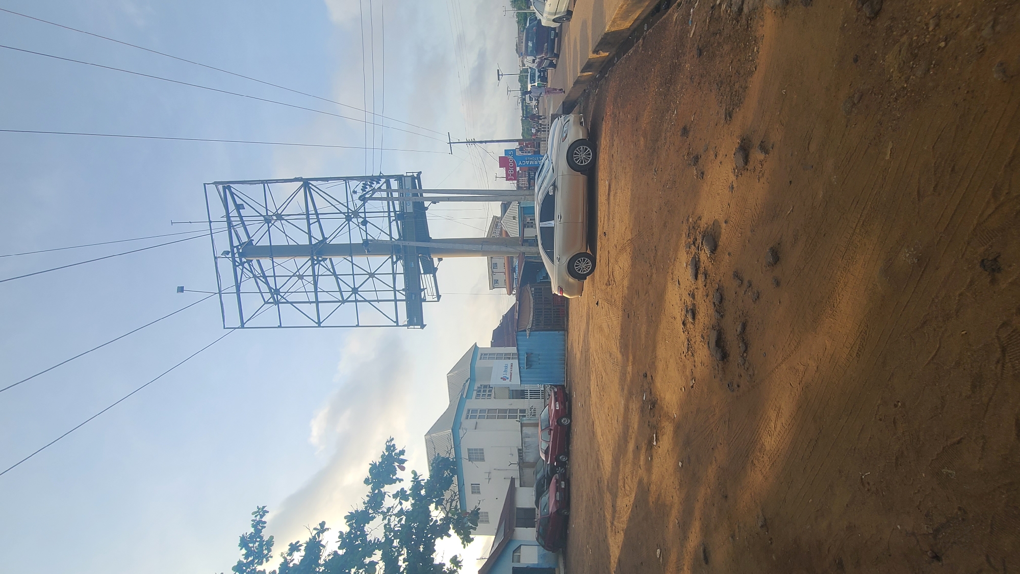 The image shows a street scene in Mandate Estate, Ilorin, Nigeria. In the foreground, there's a dirt road with tire tracks and scattered rocks. Parallel to the road, several parked cars are visible, including a light-colored sedan and multiple red sedans. Behind the cars, a building with multiple windows and a blue roof detail is present. A large, empty billboard frame stands tall, supported by a network of metal beams. Power lines stretch across the sky above the frame. The sky is partly cloudy, with visible sunshine.