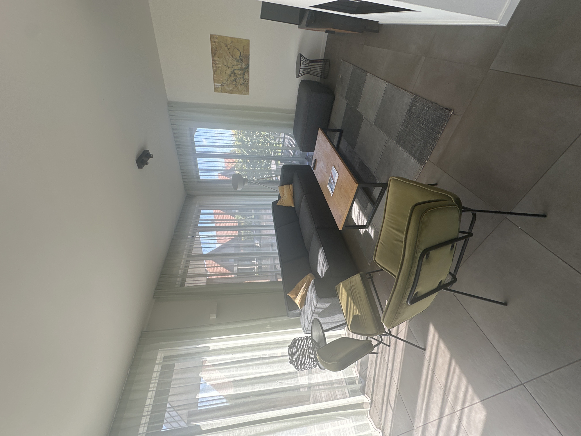 The image depicts a modern, sparsely furnished living room with large windows allowing ample natural light to stream in, creating distinct shadows on the floor. The room features a dark grey sectional sofa with a single yellow throw pillow. In front of the sofa is a light brown wooden coffee table with a dark metal frame, on which a magazine and a remote control are visible. To the left of the coffee table, two olive green upholstered chairs with thin black metal legs are positioned. A black wire basket-style side table with a round top sits near the window. Behind the sofa, a dark grey ottoman-style pouf is placed. A small, black wire side table with a rounded, open design is situated to the right of the pouf. On the wall above the pouf hangs a framed abstract painting featuring muted yellow and brown tones with tree-like imagery. To the far right, a portion of a black entertainment unit or cabinet is visible, and a black object, possibly a speaker or television, rests on top. A floor lamp with a white shade stands near the left side of the sofa, partially obscured by the curtain. The floor is tiled with large, square grey tiles. A grey rug with a patchwork design of varying grey tones is placed under the coffee table and pouf. The windows are covered by sheer white curtains that diffuse the light and reveal glimpses of residential buildings and greenery outside. The overall impression is one of a clean, contemporary, and bright living space. The image is not specifically located in Ossenzijl, Netherlands, as no identifying landmarks or context are provided within the image itself to confirm this location.