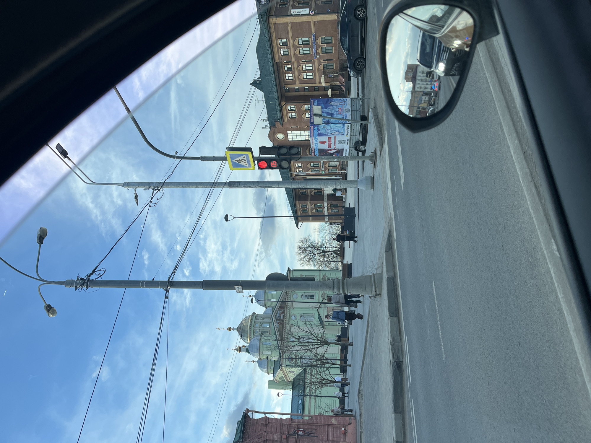 The image, taken from inside a vehicle, captures a street scene in Klin, Russia. A red traffic light is visible above a pedestrian crossing sign. A multi-story brick building is on the right side of the street. An Orthodox church with light green and silver domes is on the left. Pedestrians are walking on the sidewalk. The vehicle's side mirror shows a car and an emergency vehicle with flashing blue lights in the distance. Overhead electrical wires crisscross the sky. The sky is blue with scattered clouds.
